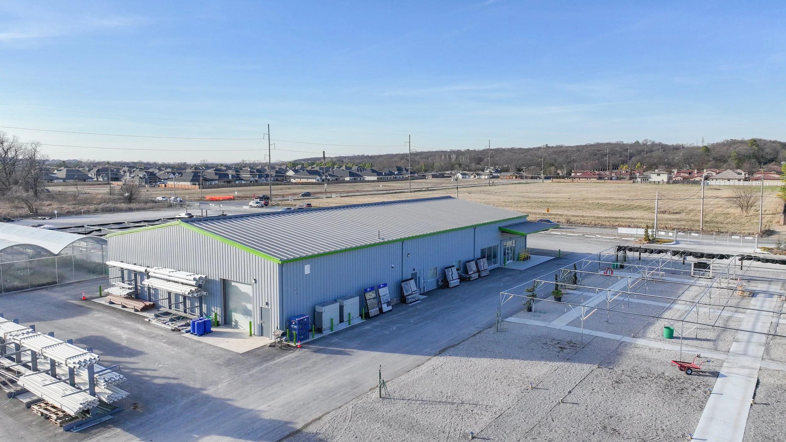 Industrial building with metal roof, outdoor displays, and gravel/soil areas, located in a semi-rural area with open fields and residential houses in the background.