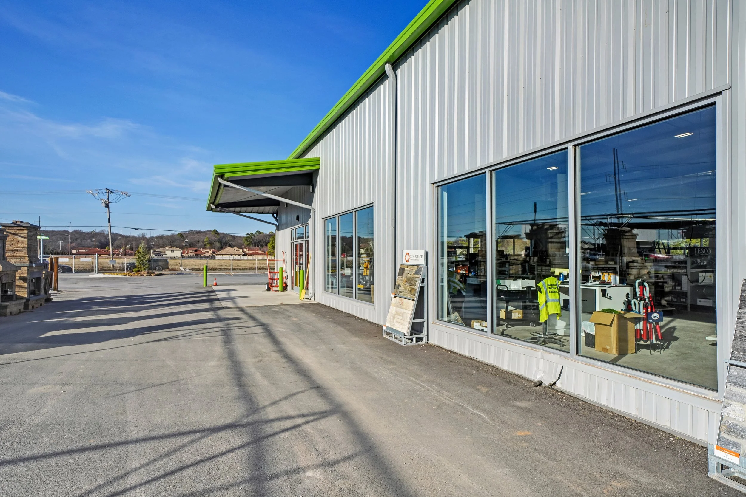 Exterior view of a hardware store with large glass windows, metal siding, and a green roof trim. The parking lot in front is mostly empty, and various tools and equipment can be seen through the windows.
