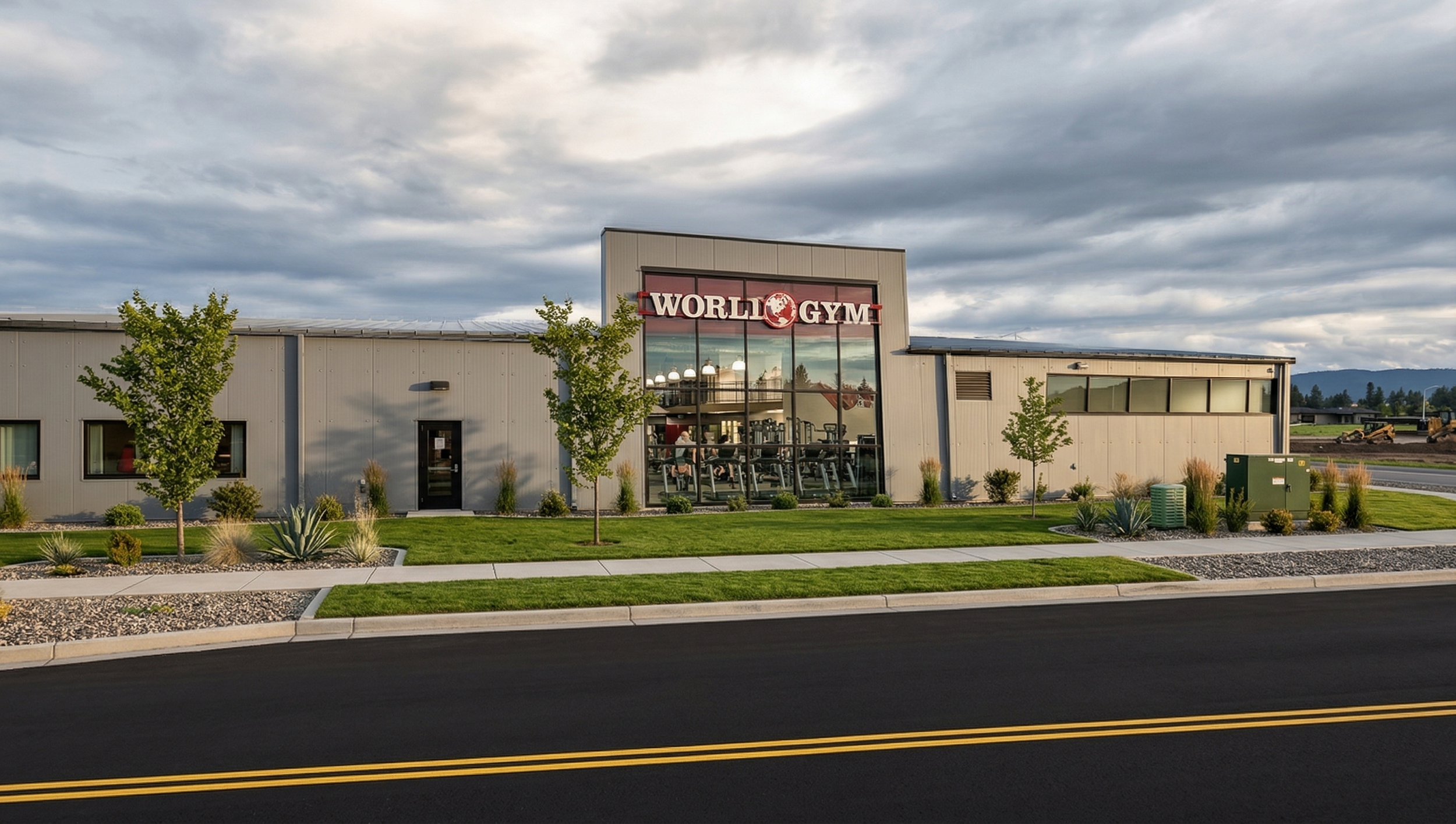 Exterior view of a fitness center named 'World Gym' with large glass windows, trees, and landscaped bushes in front, under a cloudy sky.