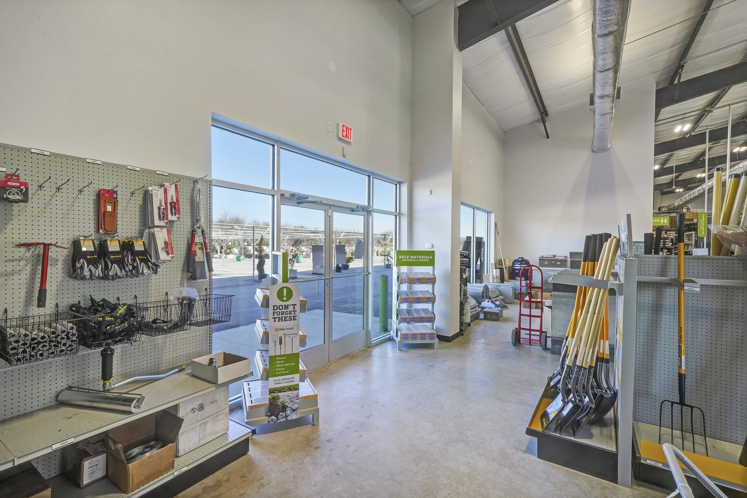 Interior of a hardware store with tools and equipment on display, including shovels, hammers, and lawn care tools, near glass doors leading outside.