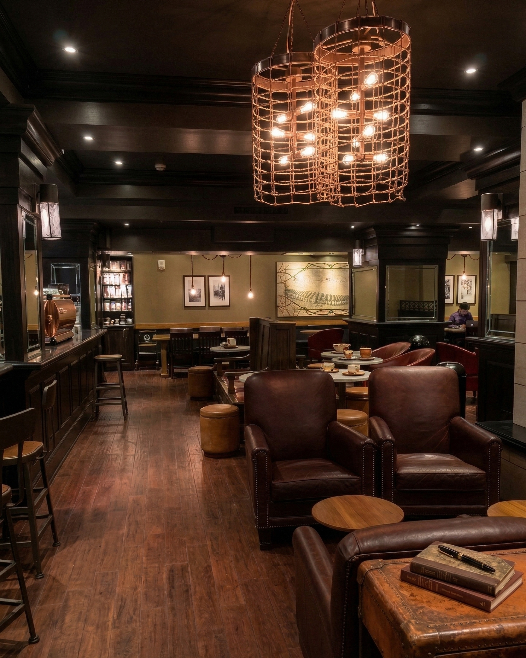 A cozy, dimly lit cafe or lounge with dark wood furniture, leather armchairs, and a modern chandelier. Tables are set with cups and saucers, and there are framed pictures and artwork on the walls.