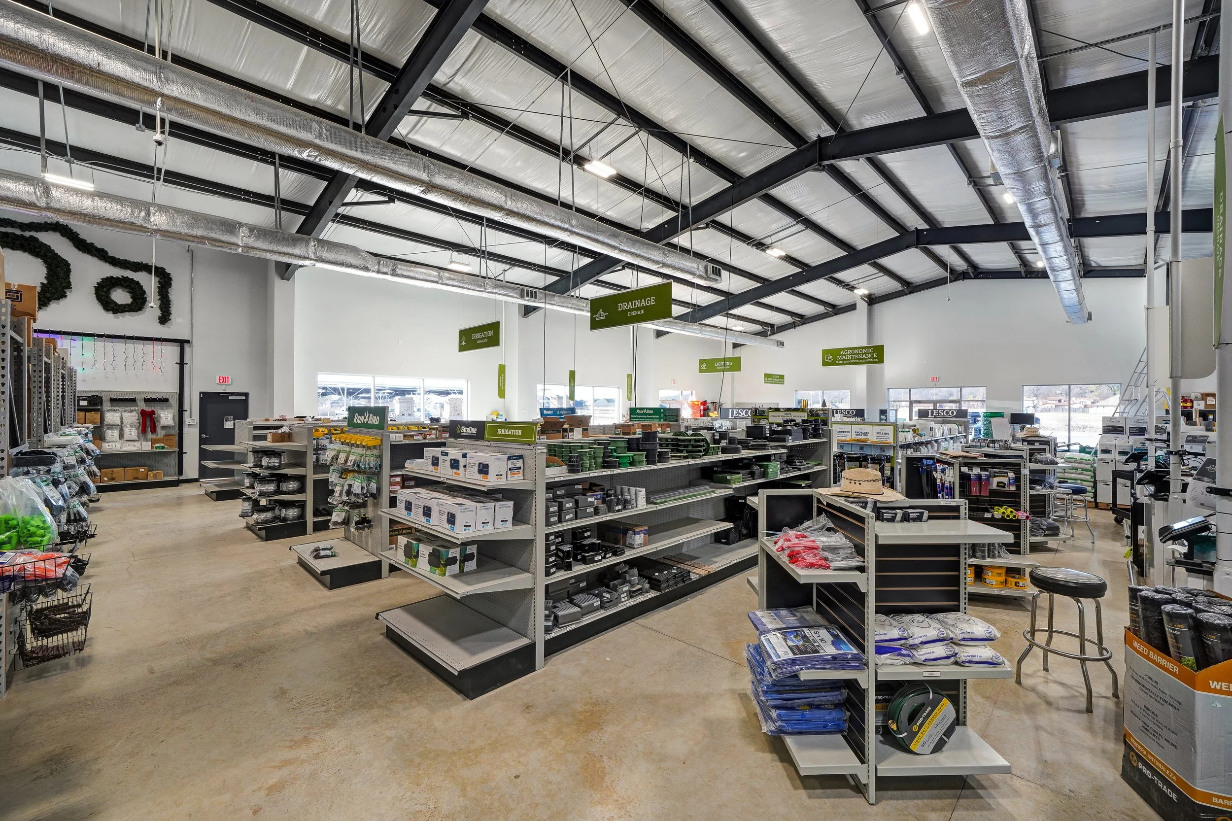Interior of a hardware store with shelves stocked with irrigation supplies, drainage products, and garden tools under a high, industrial ceiling with exposed ductwork and large windows.