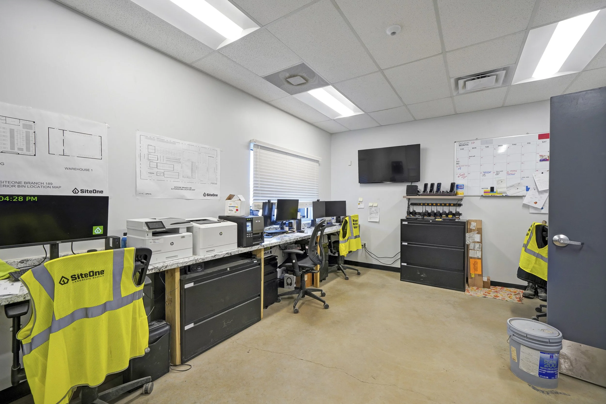 An office with white walls, ceiling with fluorescent lights, a window with blinds, multiple desks with computers, printers, and office supplies. Yellow safety vests with the SiteOne logo hang on chairs. There are black filing cabinets, a large wall-m