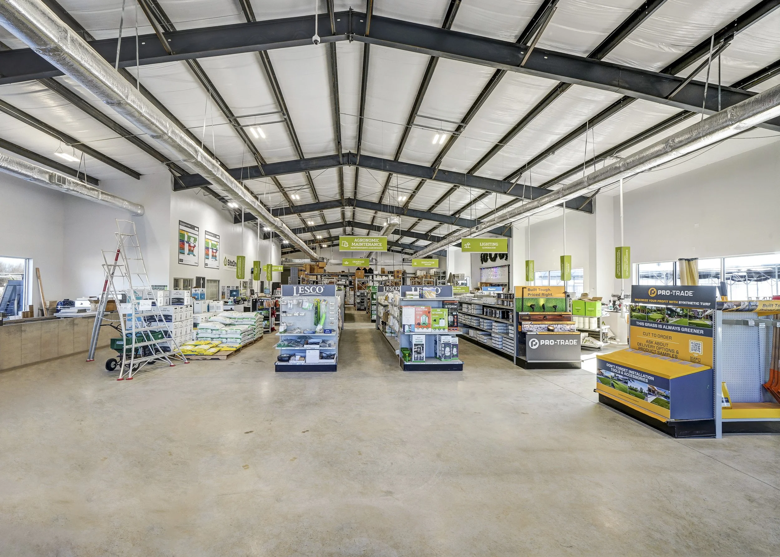 Interior of a hardware store with shelves stocked with gardening, lighting, and maintenance supplies, and a large open floor space.