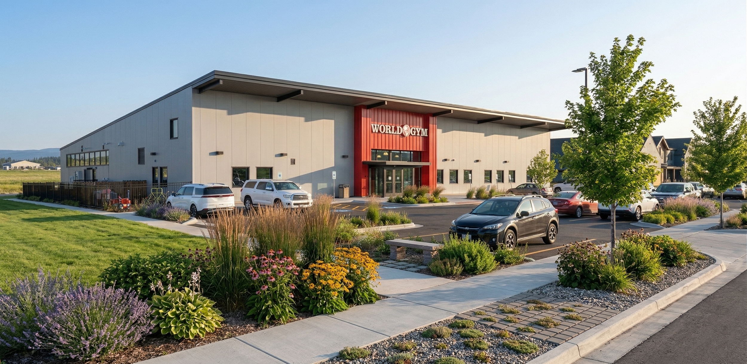 Exterior view of a modern gym building labeled 'World Gym' with a parking lot and landscaped area with flowers and trees.