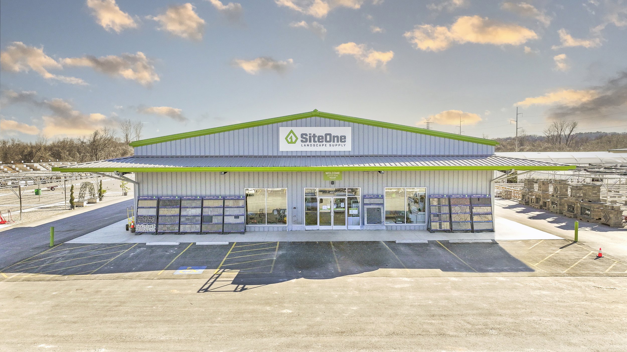 A landscape view of a StoreOne Landscape Supply building with a green roof and white exterior, surrounded by outdoor displays of landscaping materials, parking spaces, and a clear sky with clouds.