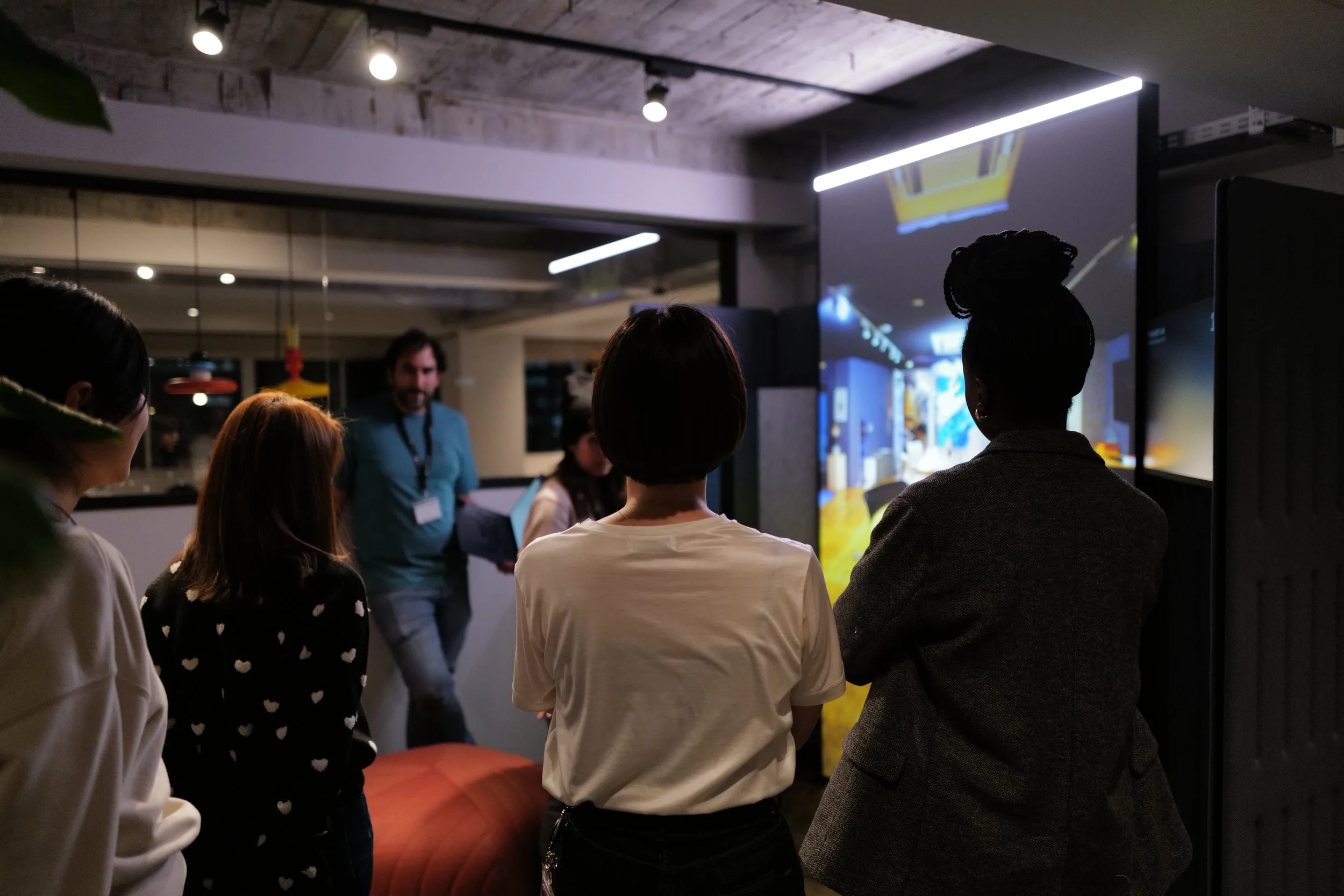 A group of people in an office setting, focusing on a digital screen displaying a virtual interior design. The room has modern lighting and a relaxed, collaborative atmosphere.