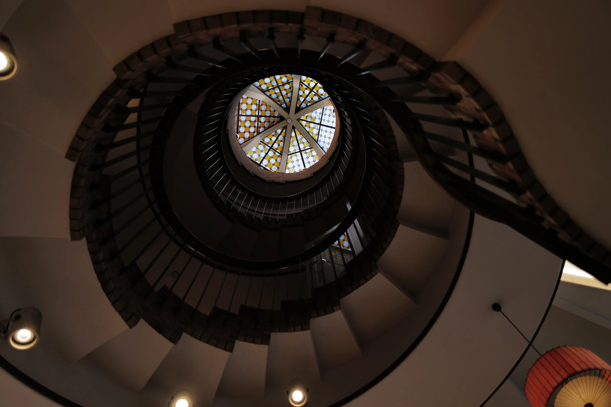 Spiral staircase leading up to a stained glass ceiling with geometric patterns, viewed from below, in a dimly lit interior.