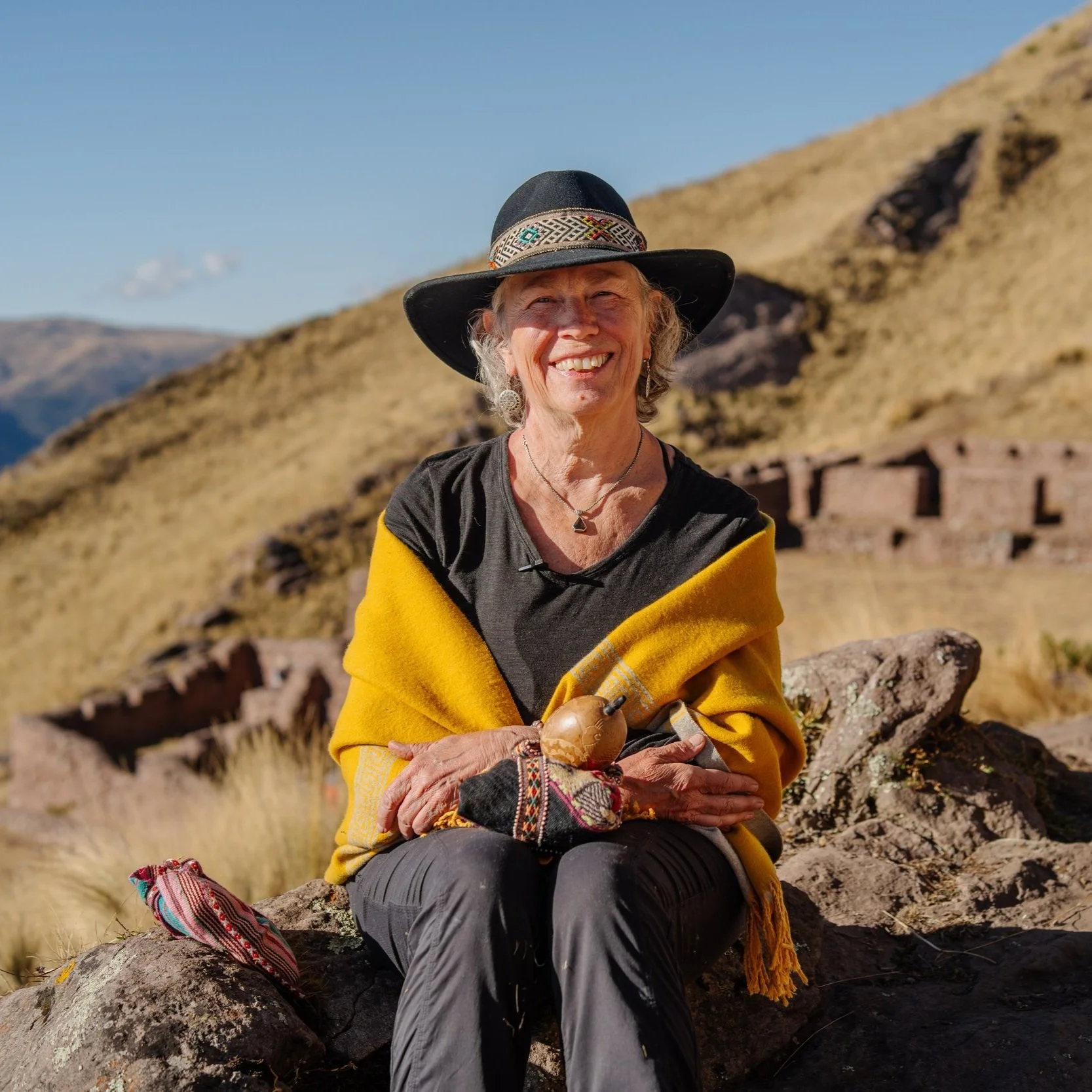 Woman in yellow shawl and black hat sitting on a rock with a scenic mountain backdrop.
