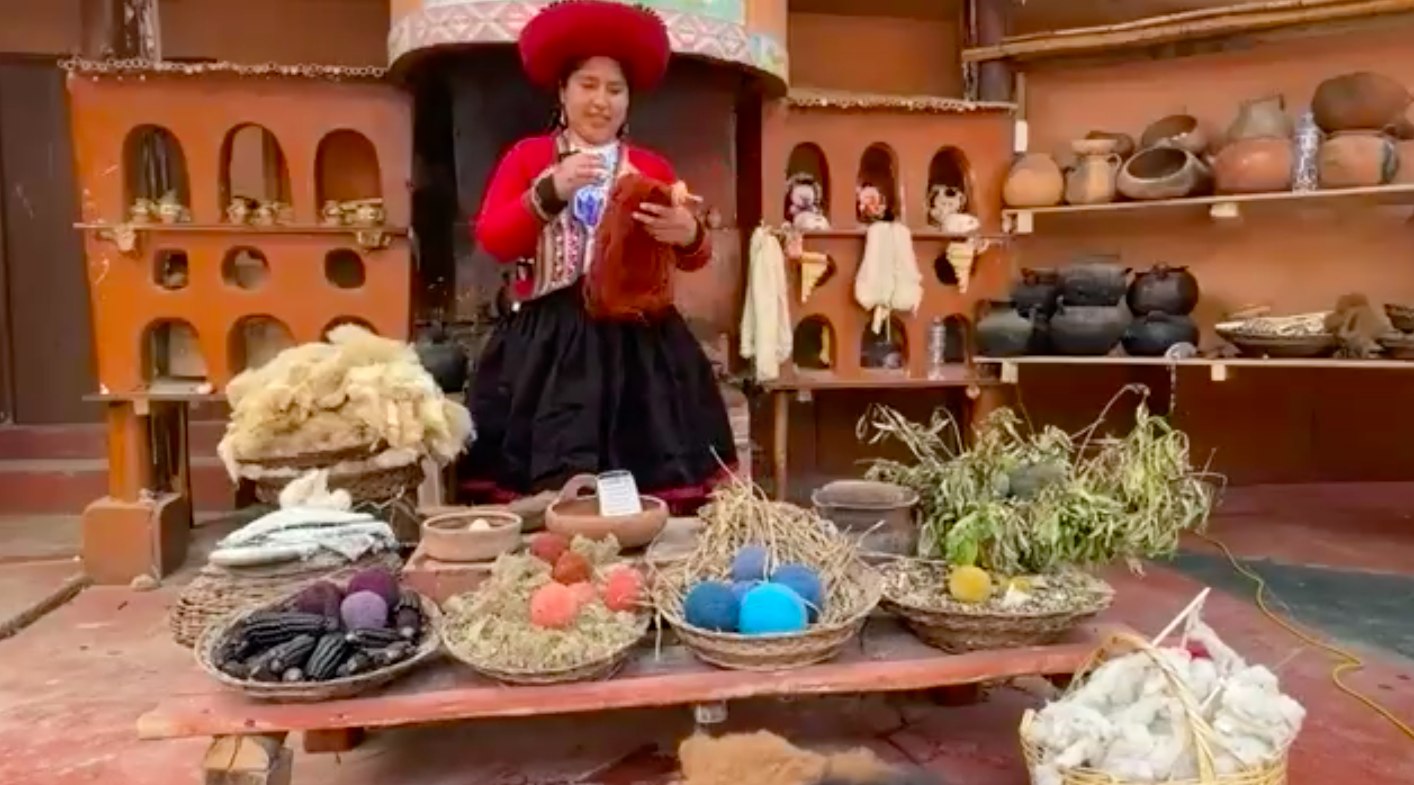 Woman in traditional clothing demonstrating weaving materials, including dyed wool and plants, in a traditional setting with pottery and textiles on display.