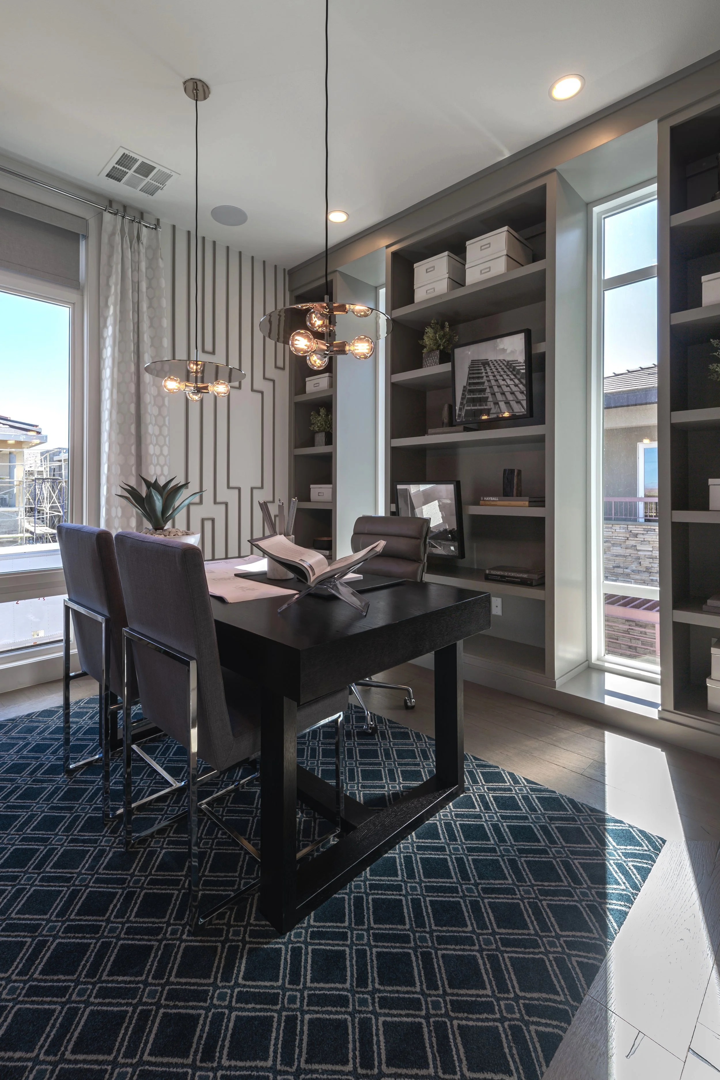 Modern home office with a dark wooden desk, two chairs, open shelving with decor, geometric pendant lights, and a patterned rug.