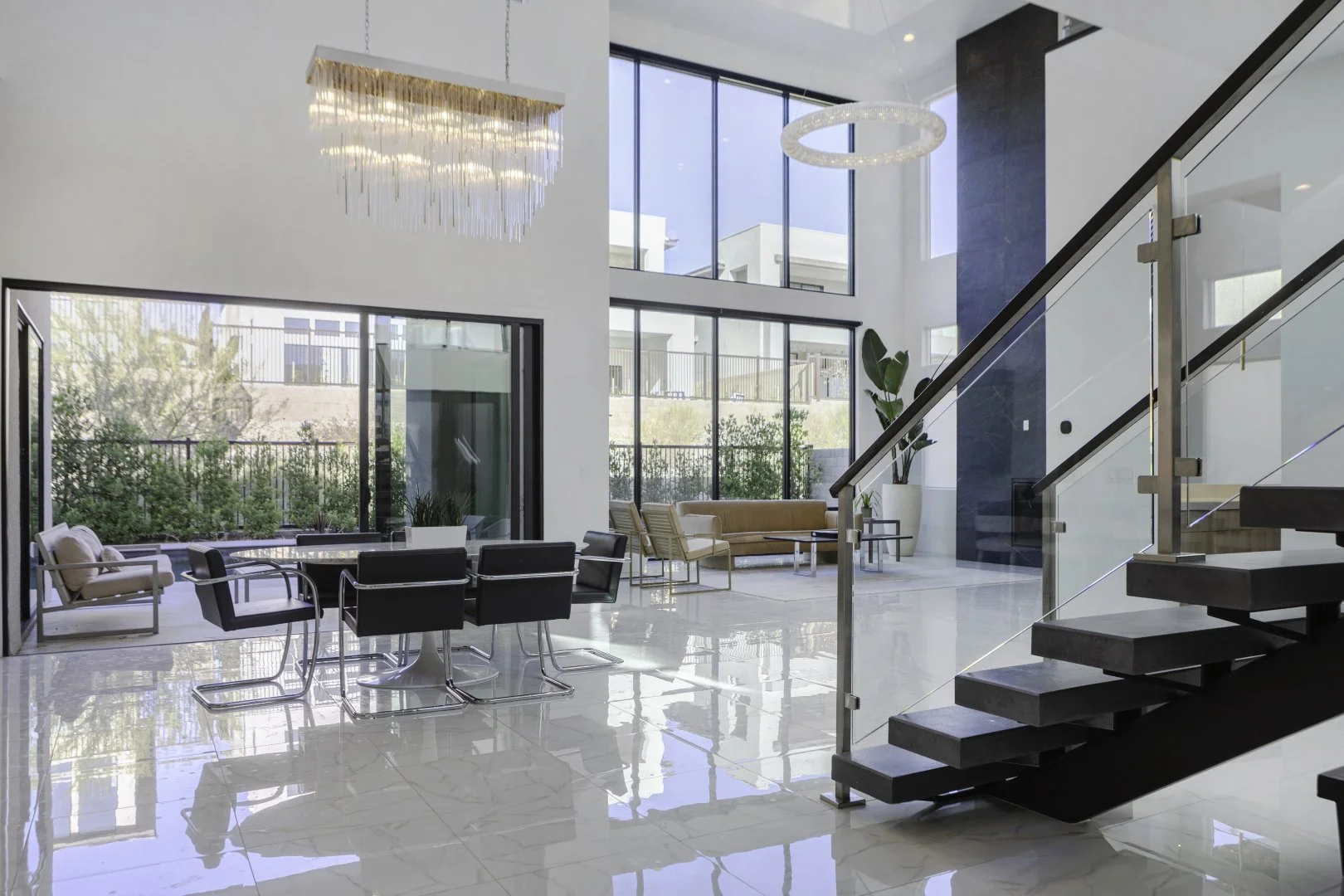 Modern living room interior with staircase, large windows, and contemporary furniture; featuring a chandelier and glass railing.