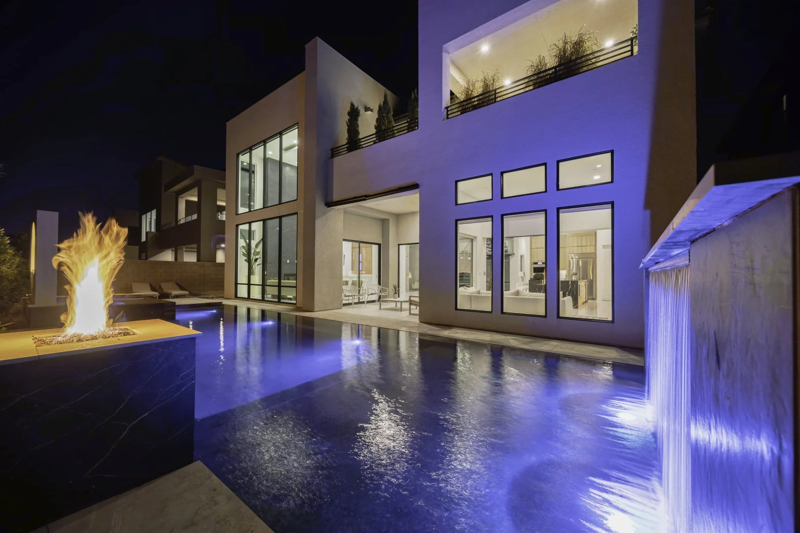 Modern luxury house exterior at night, featuring large windows, a lit swimming pool, fire pit, and waterfall.
