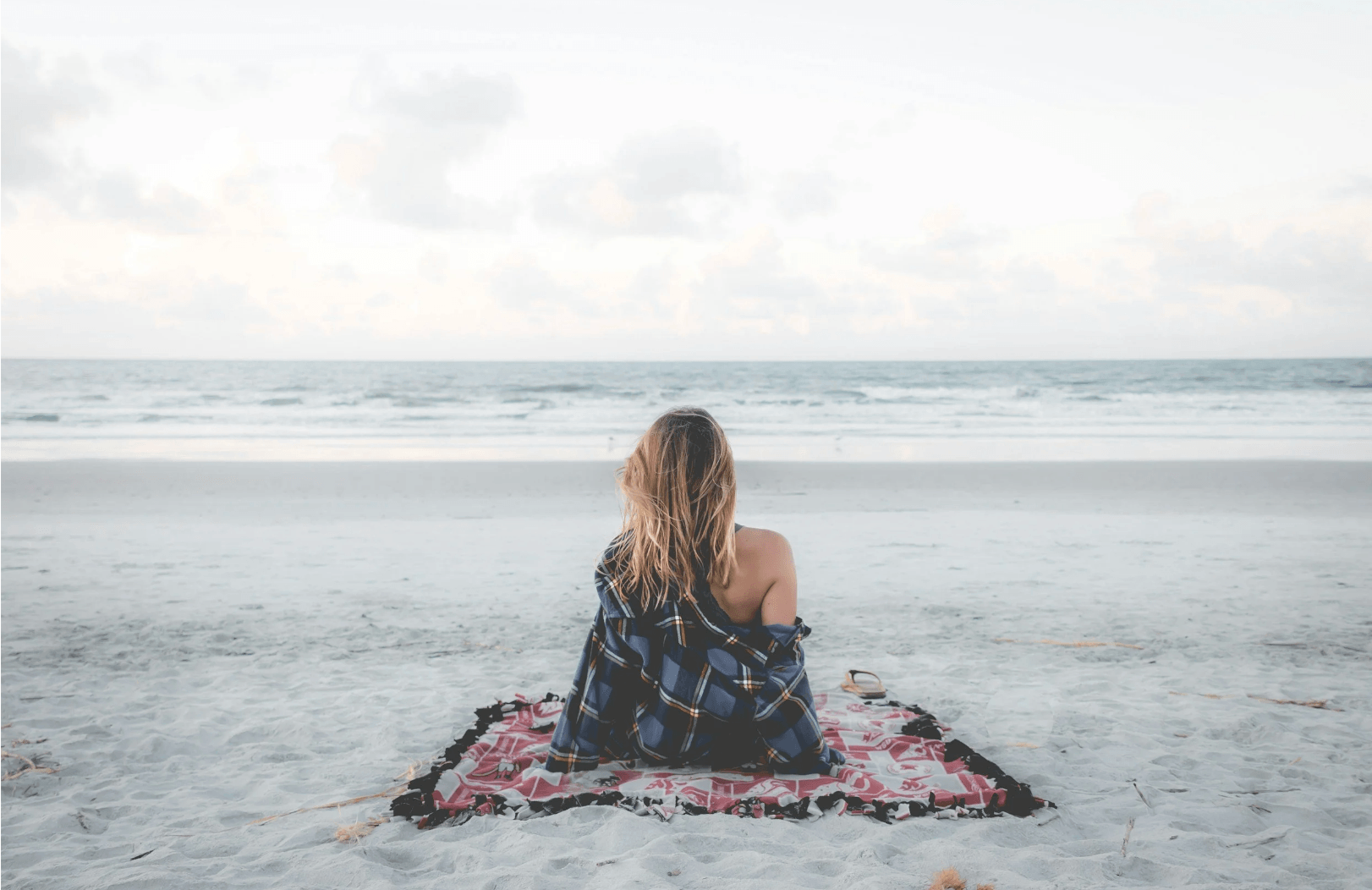 Person sitting on a blanket at the beach, facing the ocean, with a plaid shirt draped on their shoulders.
