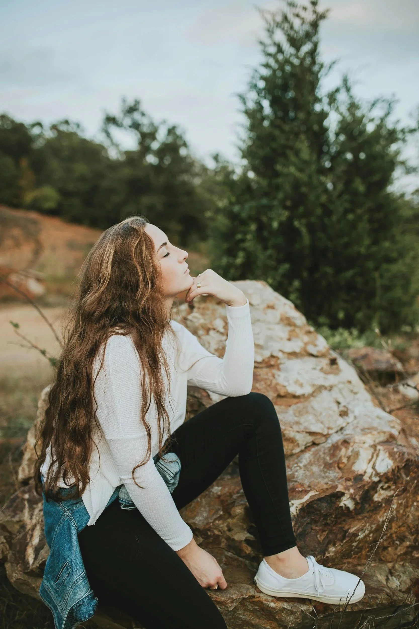 Woman with long curly hair sitting on rocks outdoors, gazing thoughtfully to the side with trees and sky in the background.