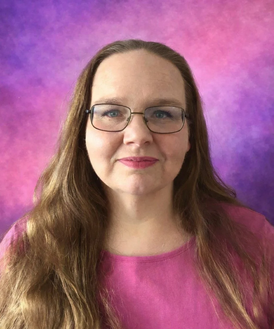 A woman with long brown hair, glasses, and pink lipstick standing in front of a pink and purple background, wearing a pink shirt.