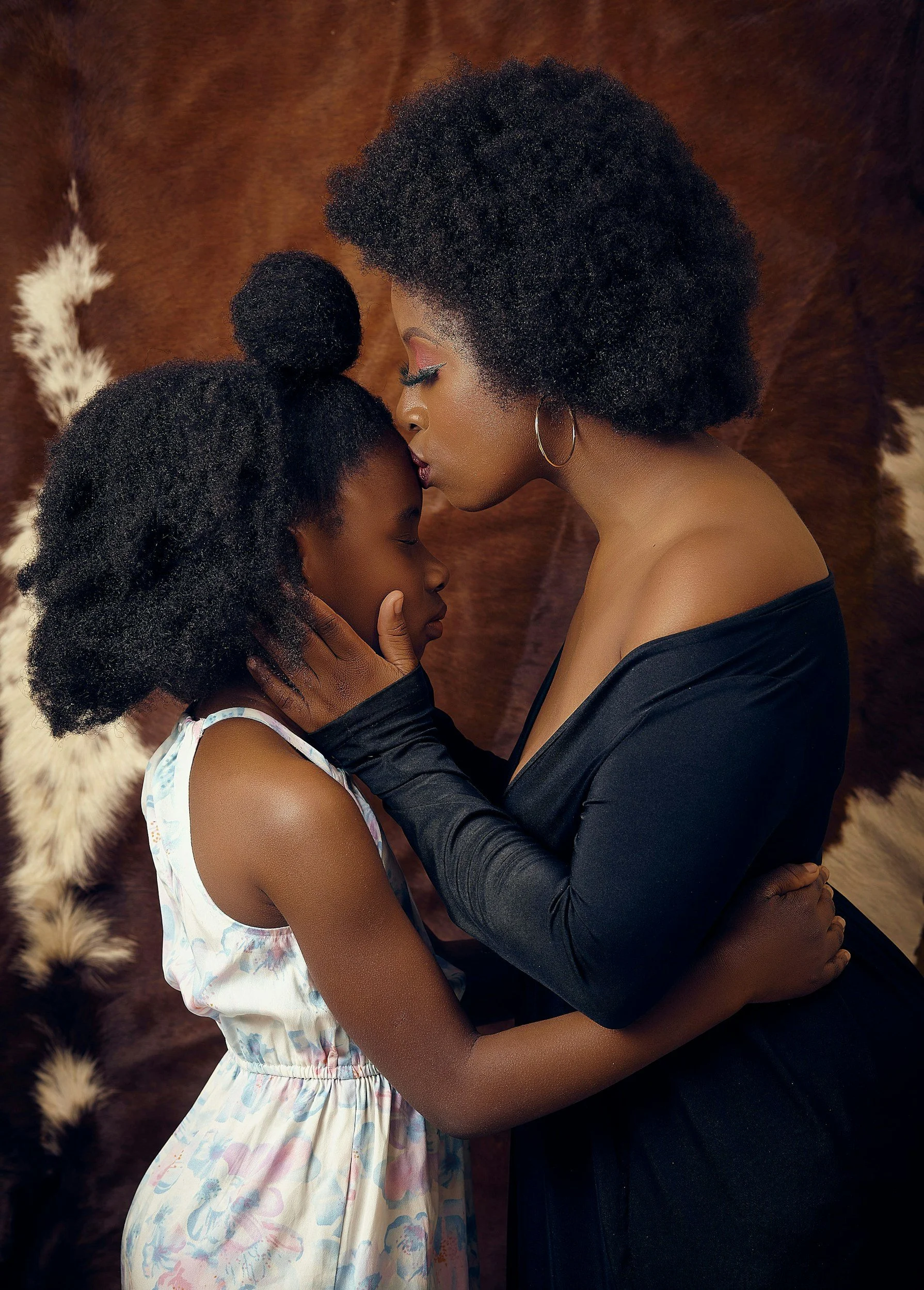 An adult woman and a young girl share a tender moment, with the woman kissing the girl's forehead. They are holding each other closely, against a furry, animal-print background. The woman has curly hair, wears hoop earrings, and a black off-shoulder top, while the girl has curly hair styled in different buns and wears a sleeveless dress with a colorful pattern.