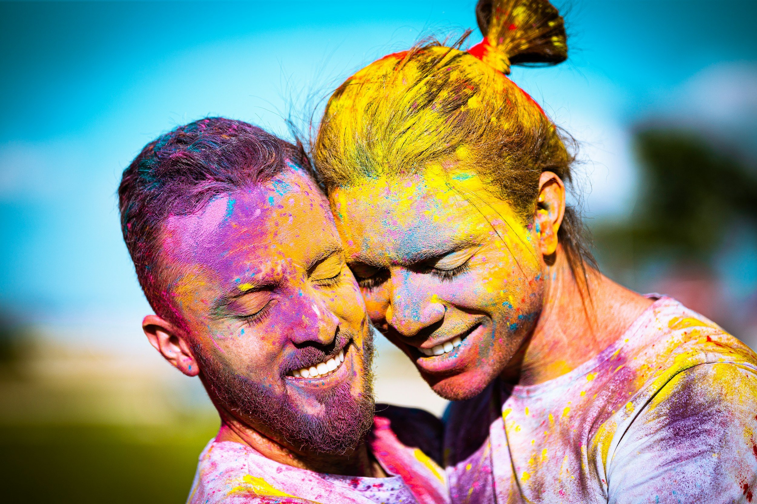Two people happily embracing, covered in colorful powder, celebrating outdoors, with a clear blue sky in the background.
