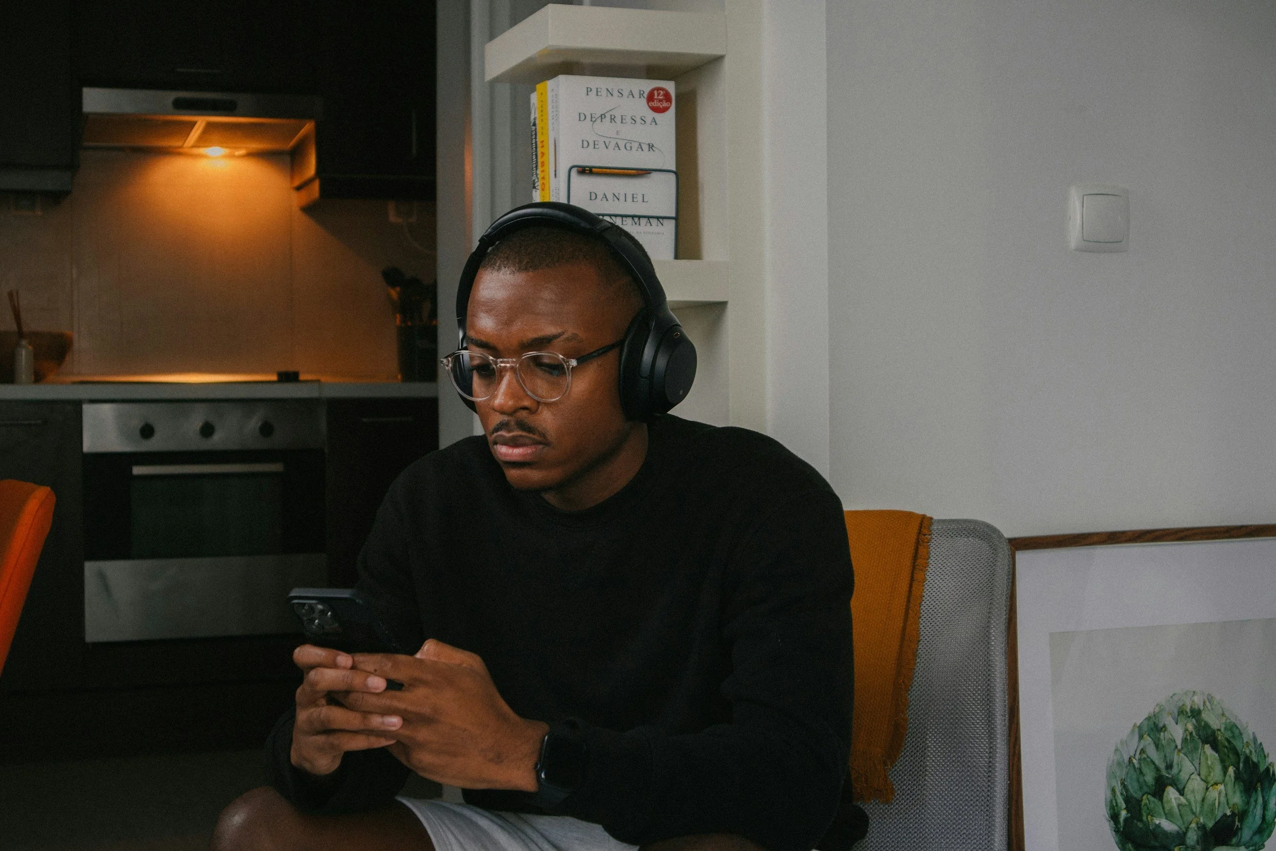 A young man with glasses and headphones sitting on a chair, looking at his phone, in a room with a modern kitchen in the background.