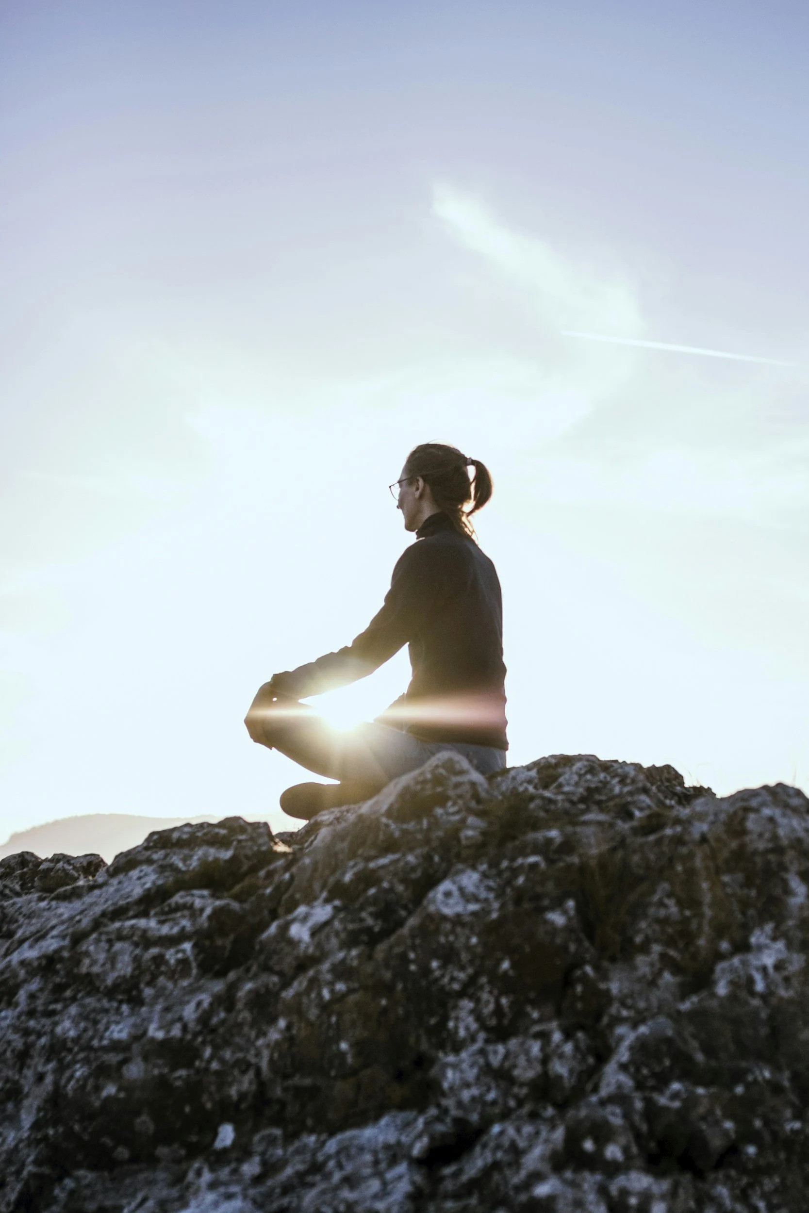 Person practicing meditation outdoors on rocky terrain at sunset with visible sun rays, mountain in background, and sky with clouds.