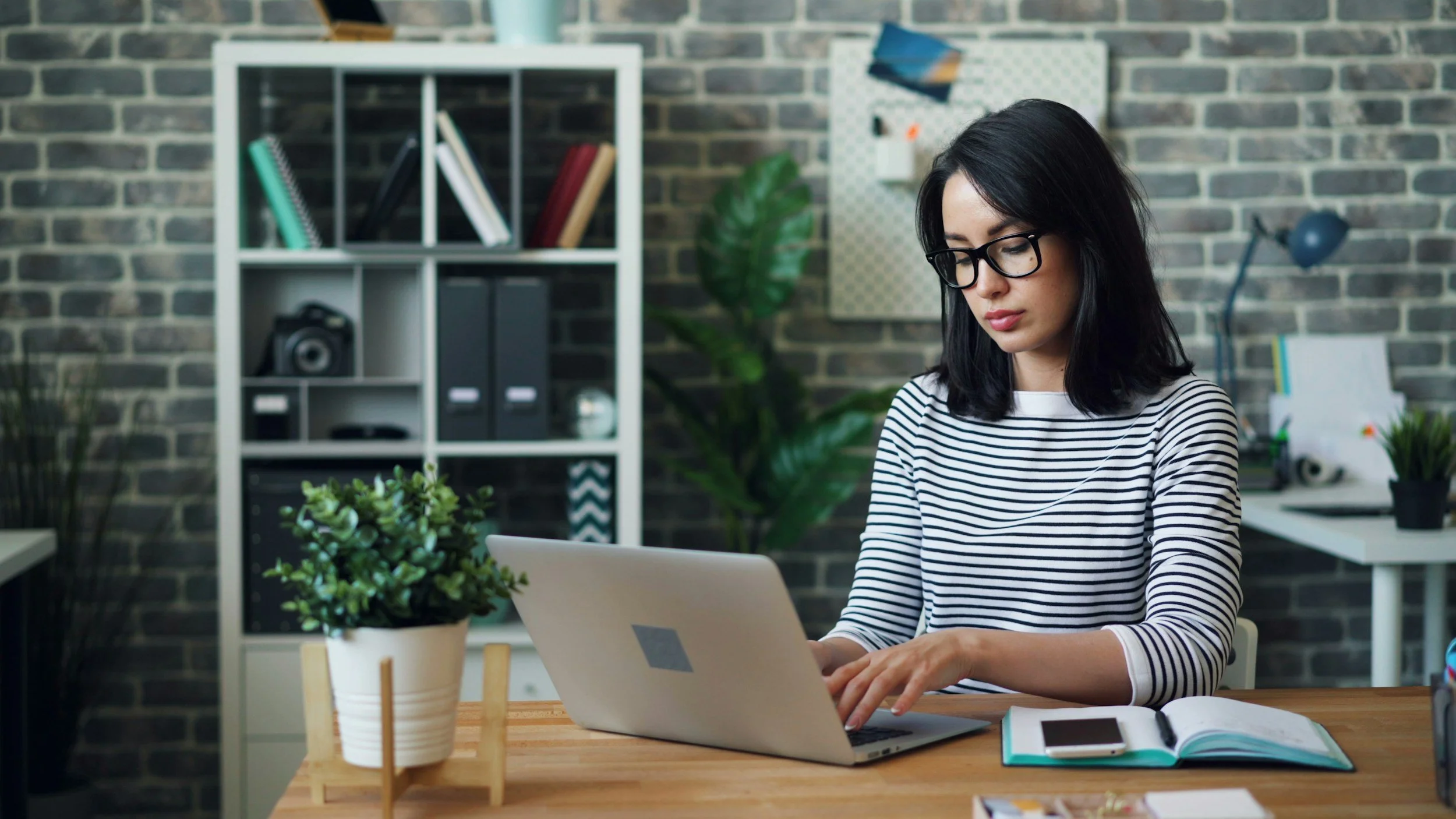 A woman with black hair and glasses working on a laptop at a wooden desk, with a potted plant, open notebook, and smartphone nearby, in a modern office with brick walls and bookshelf.