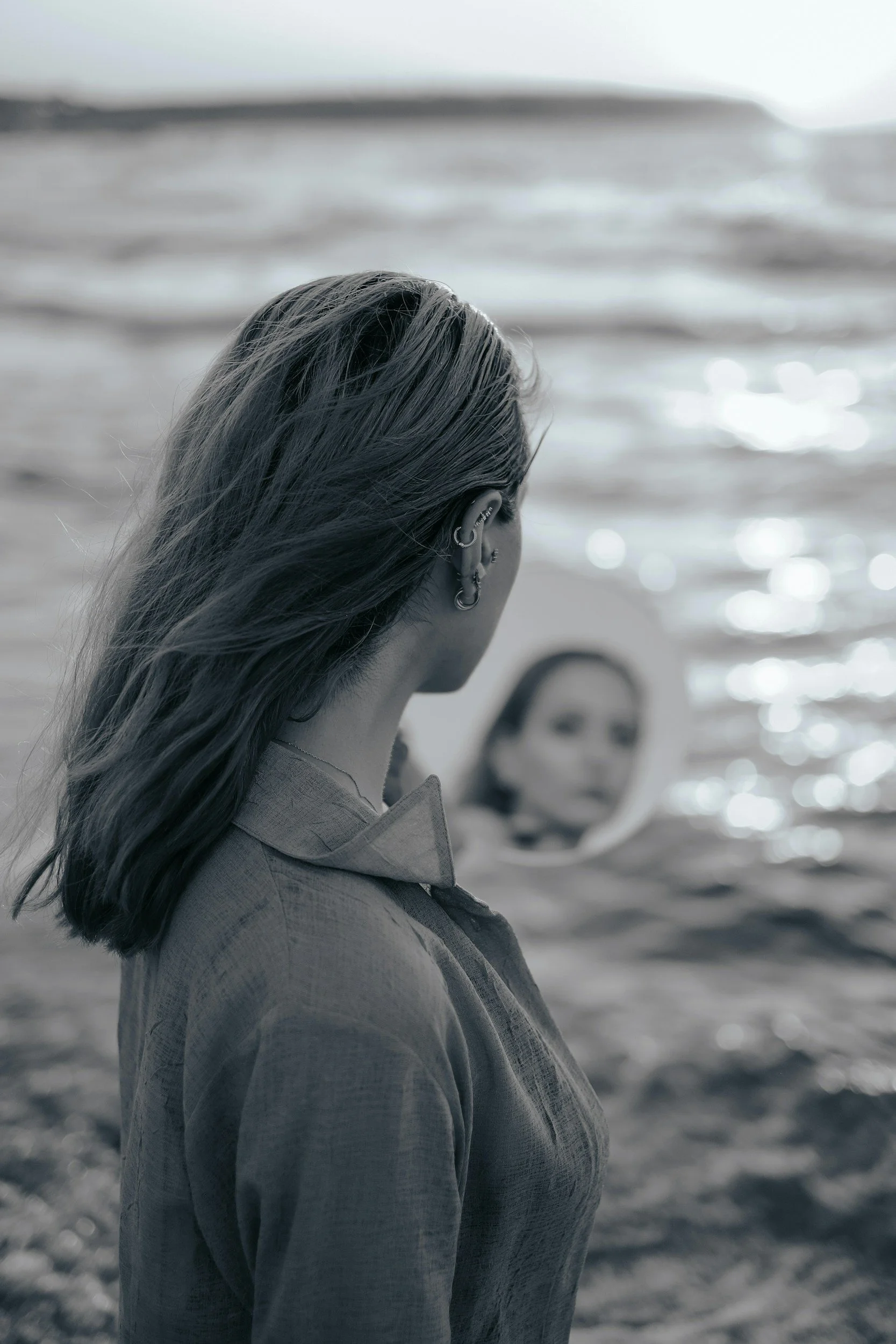 A woman standing on the beach looks into a mirror showing her face, with ocean waves in the background.