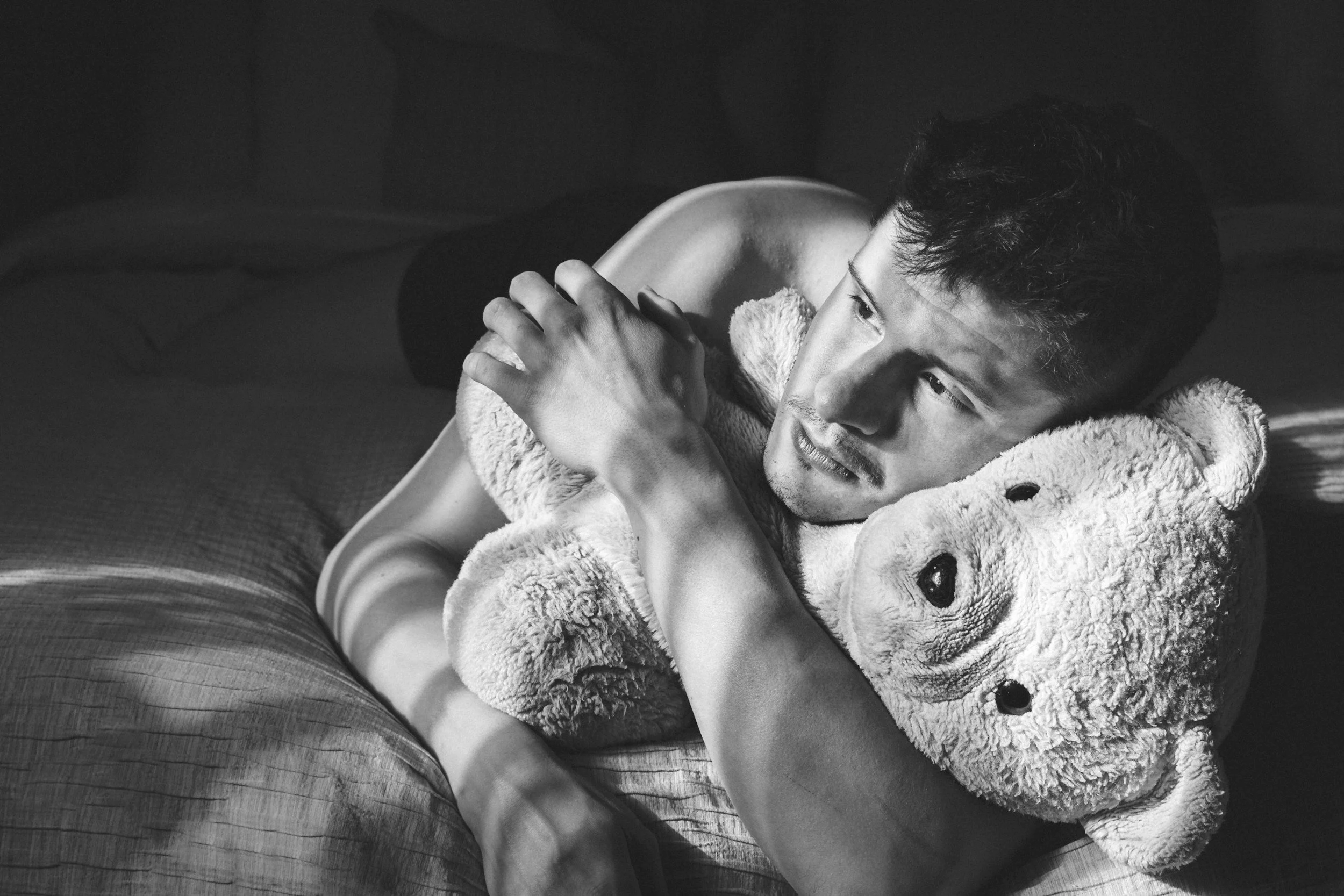 A young man lying on a bed, hugging a teddy bear, looking pensively away from the camera in black and white.