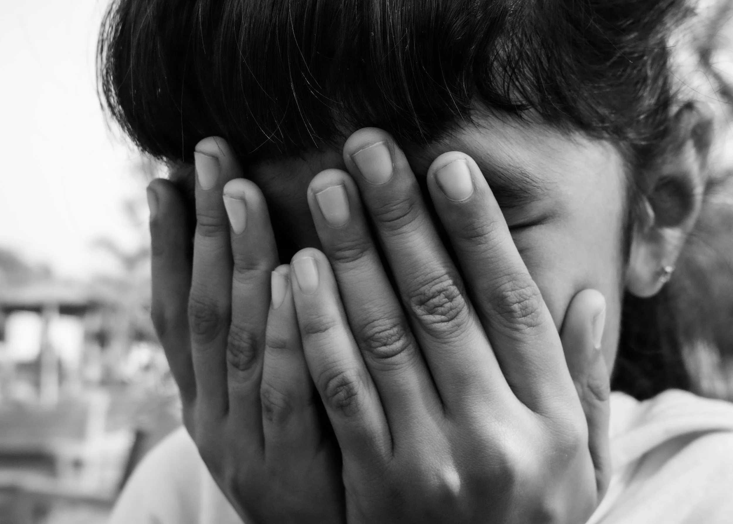 Black and white photo of a person covering their face with both hands, eyes closed, outdoors.