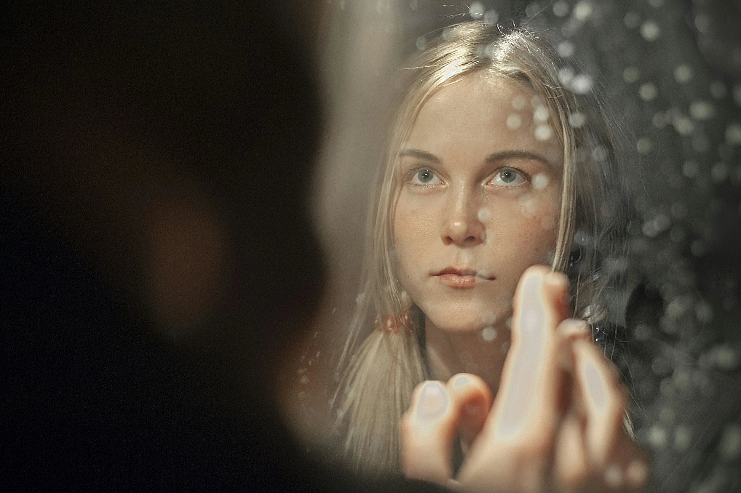 Close-up of a young woman with blonde hair and blue eyes, looking at her reflection in a mirror with water droplets on it. She is touching the mirror with her finger.