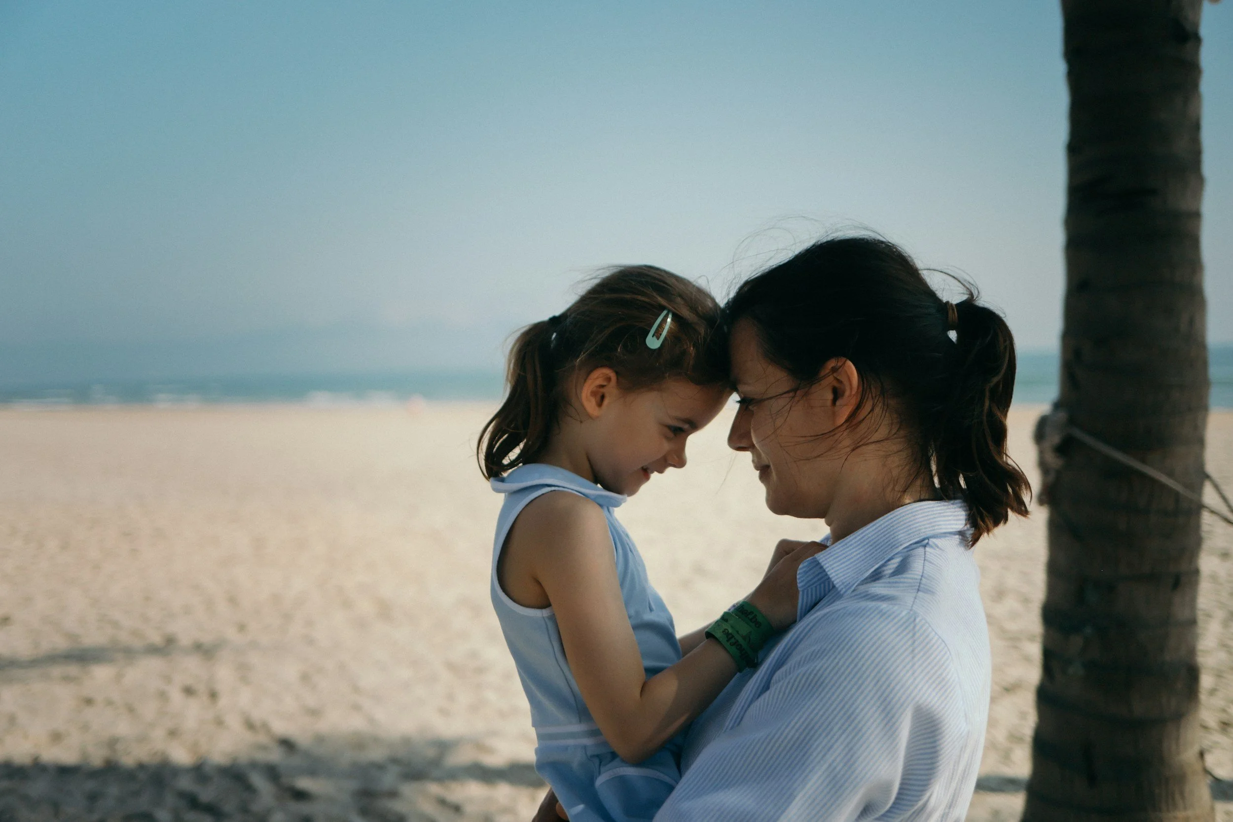 A woman and a young girl are smiling and touching foreheads on a beach with sand, ocean, and a palm tree in the background.
