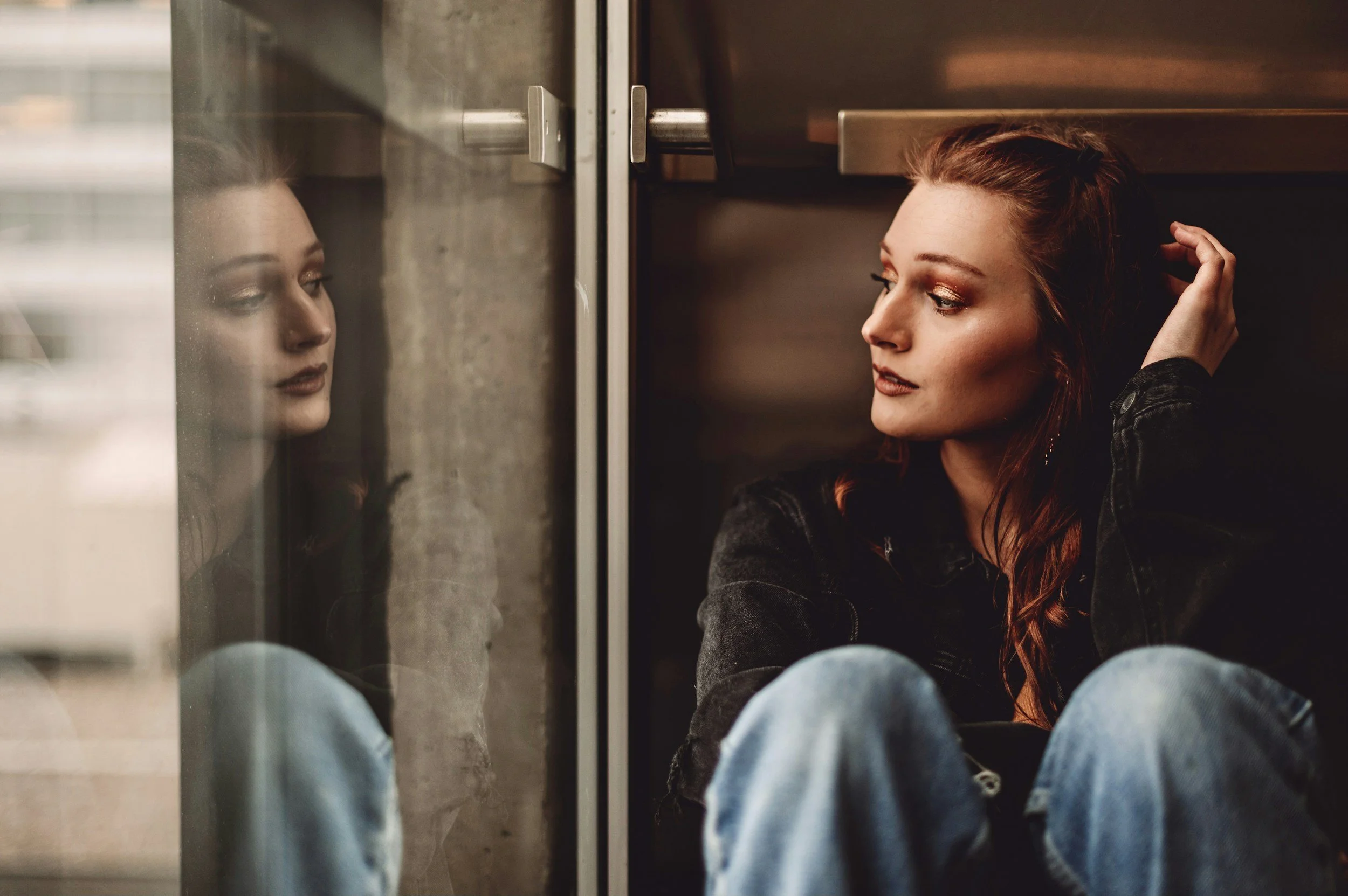 Young woman with red hair sitting on the floor in a corner, leaning against a wall, looking thoughtfully at her reflection on a glass surface.