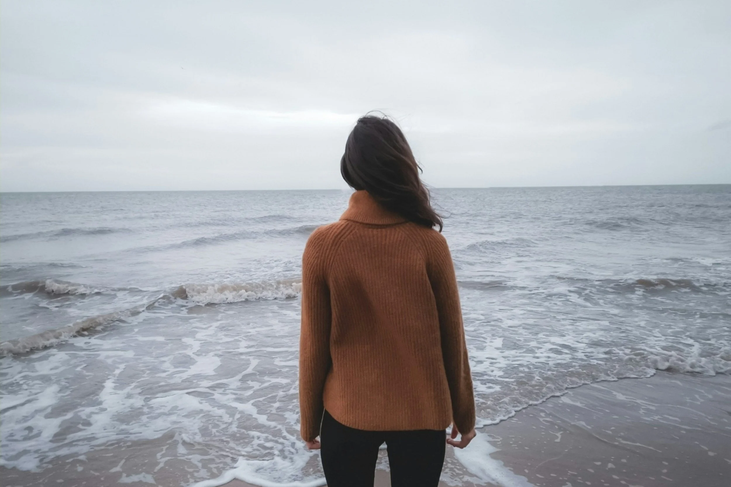 A woman in a brown sweater standing on a beach facing the ocean on a cloudy day.
