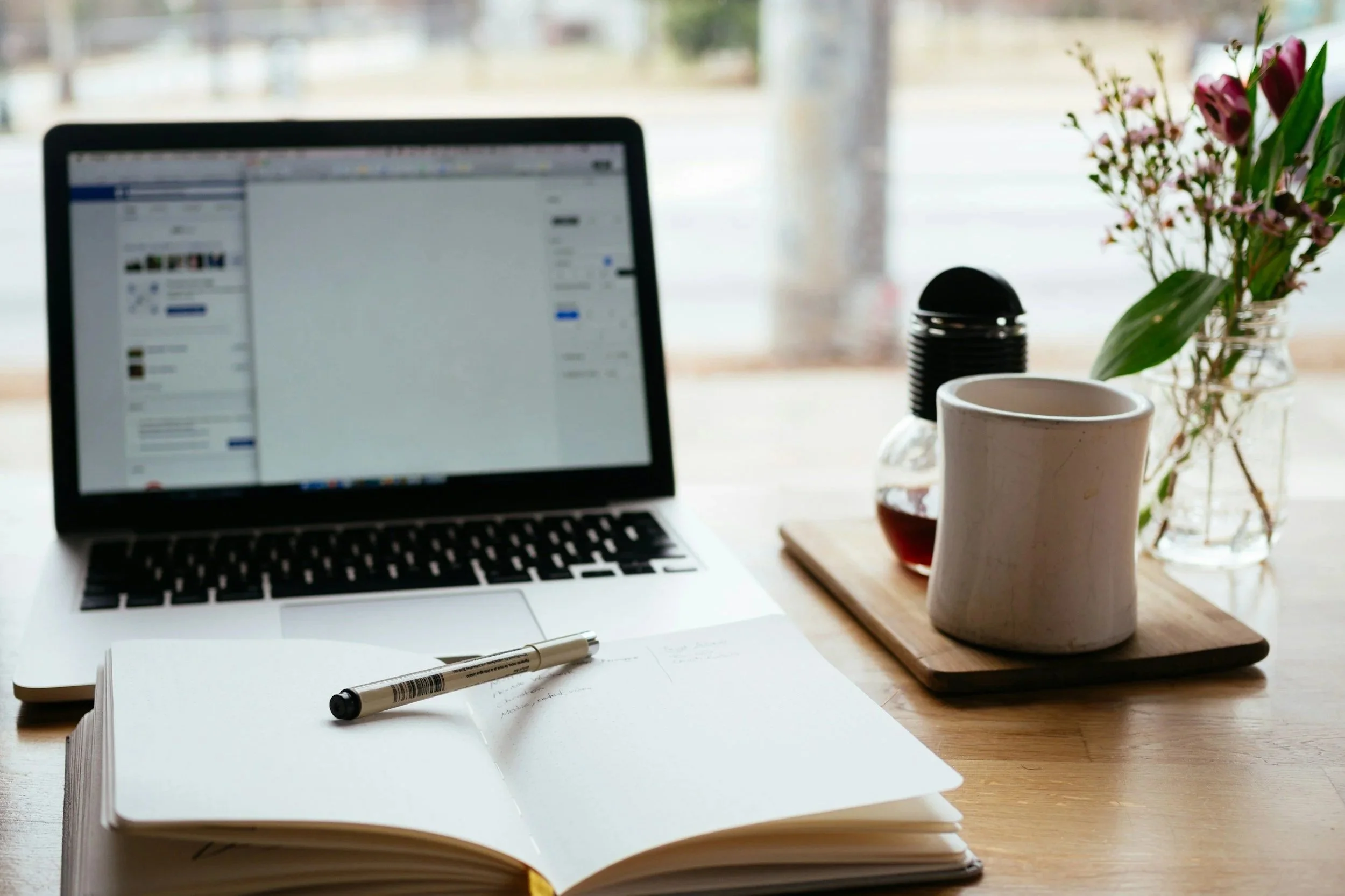 A workspace with an open laptop displaying a social media page, a closed notebook with a pen on top, a wooden tray holding a mug, a small bottle, and a jar of pink flowers, all on a wooden table near a window.