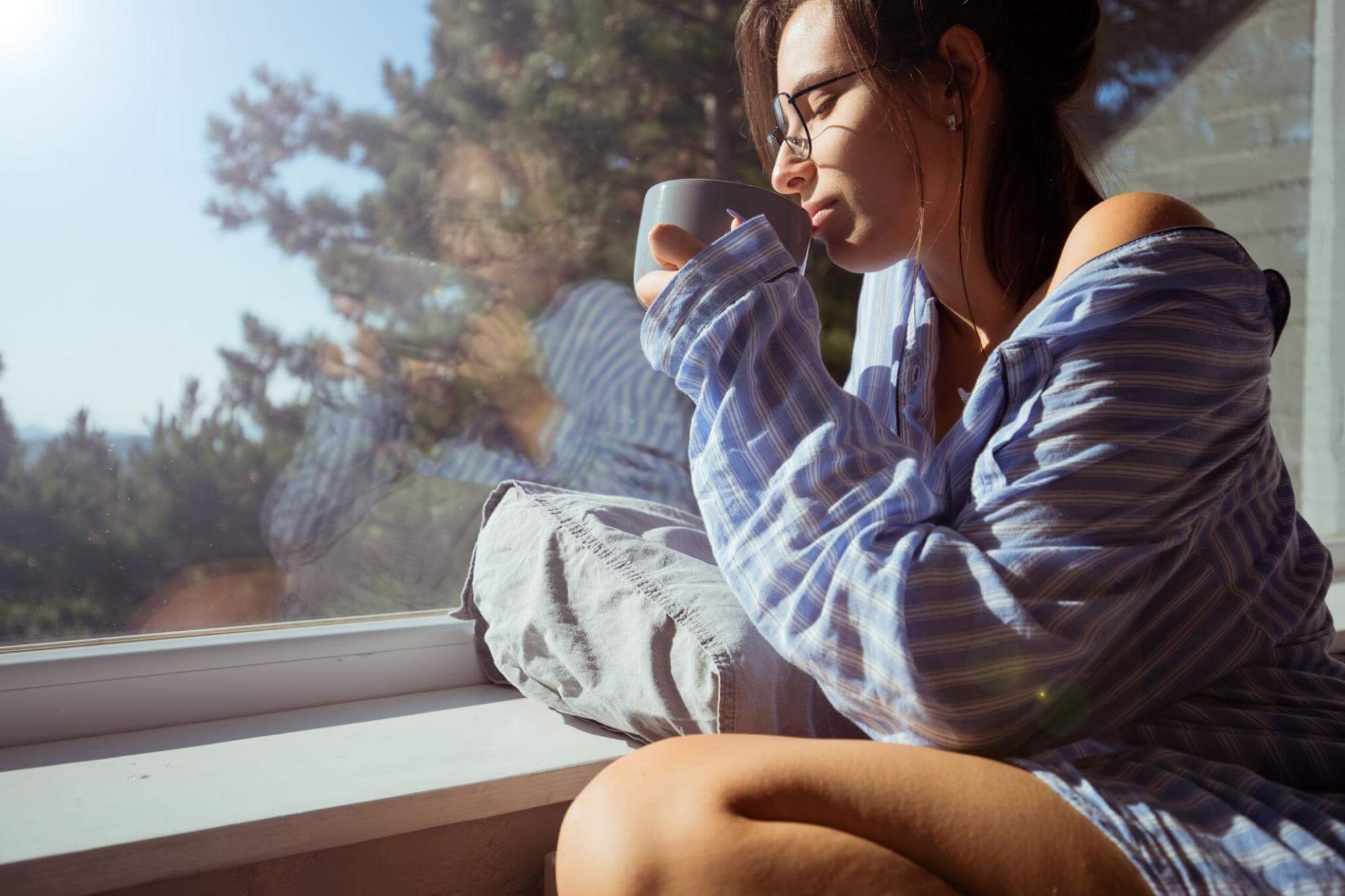 A young woman with glasses and long hair sitting by a window, holding a mug and enjoying a hot drink, with trees visible outside.