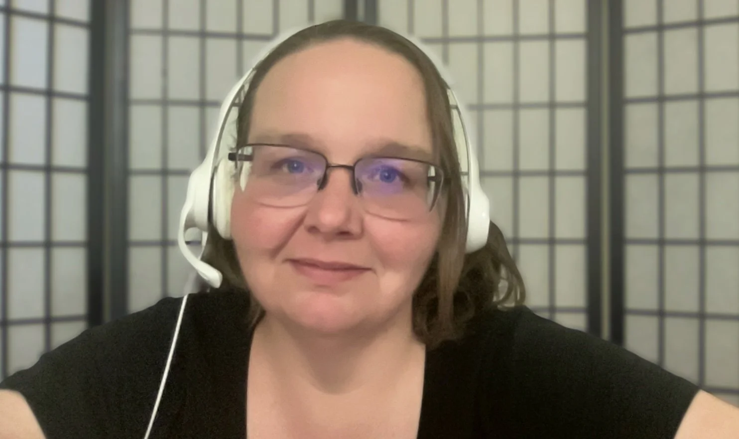 A woman with glasses and shoulder-length brown hair wearing a headset, sitting in front of a shoji screen