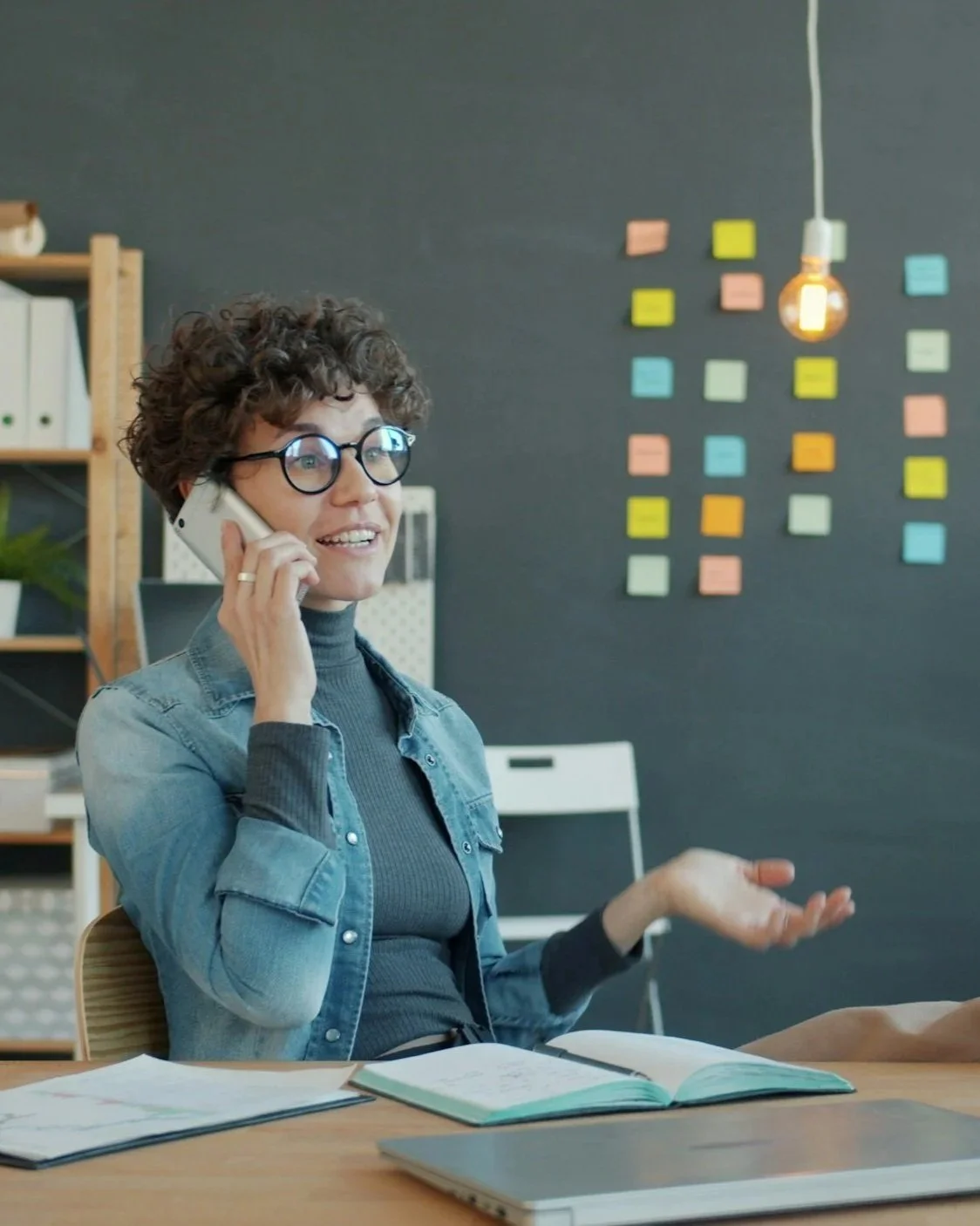 A woman with curly hair, glasses, and a denim jacket talking on the phone inside an office, with colorful sticky notes on the black wall behind her.
