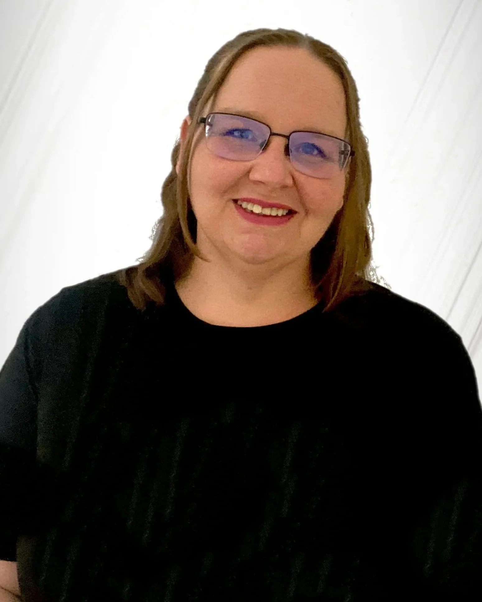 A woman smiling, wearing glasses and a black top, standing in front of a white wall.
