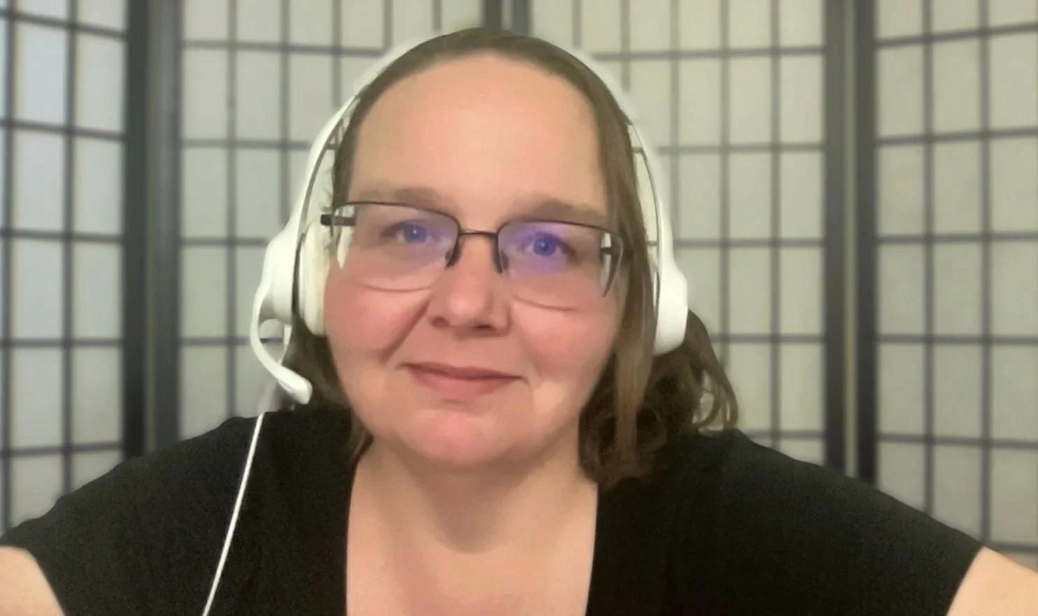 A woman with shoulder-length brown hair, glasses, and a black shirt, wearing a headset with a microphone, smiling at the camera with a black and beige folding screen in the background.