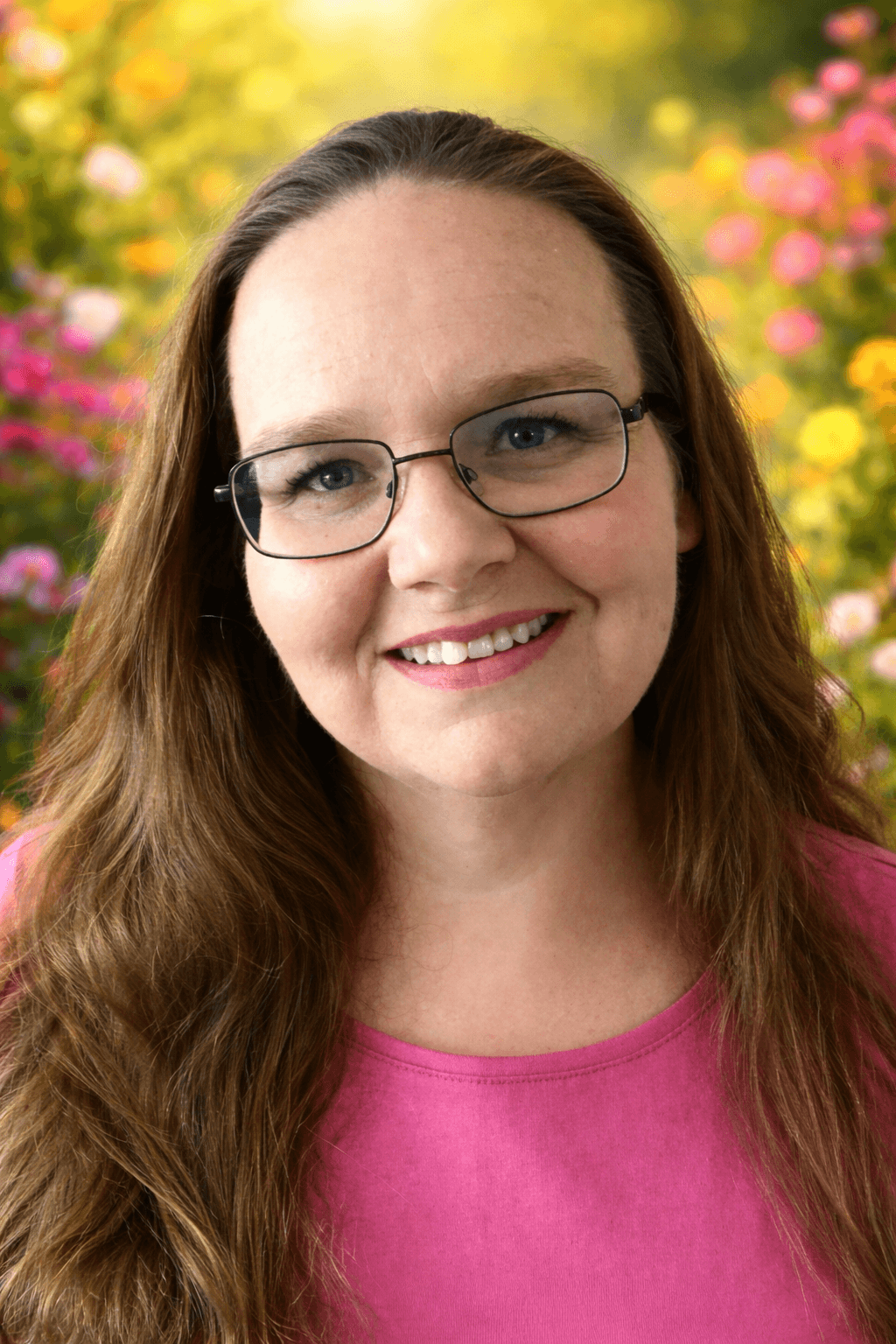 A woman, Cindy Lee Collins, Trauma therapist,  with long dark blonde hair, glasses, and a pink top smiling with a colorful, floral garden background.