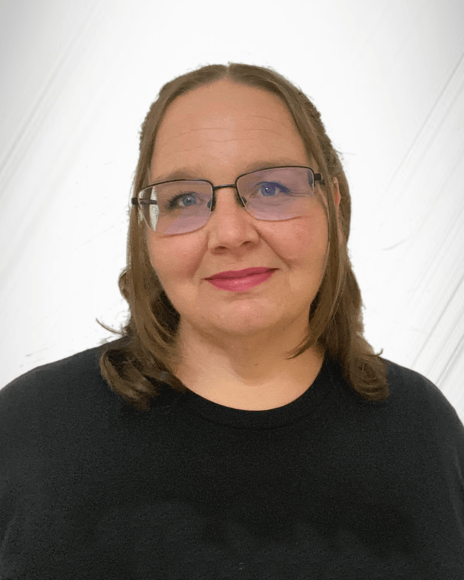 Close-up photo of a woman with fair skin, medium brown hair, wearing glasses and a black top, smiling against a white wall background.