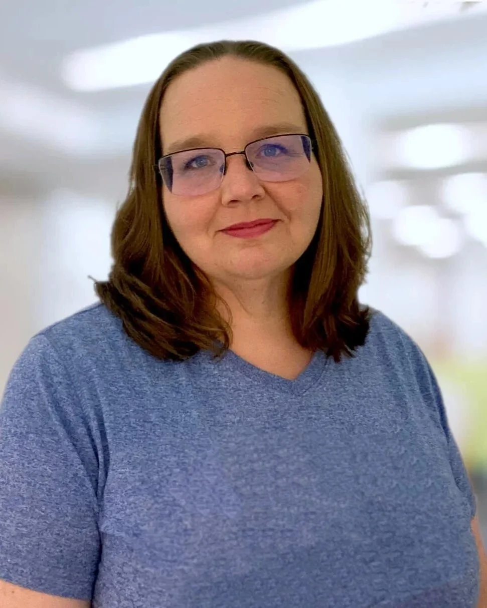 A woman with shoulder-length brown hair, wearing glasses and a blue shirt, standing in a bright indoor setting.