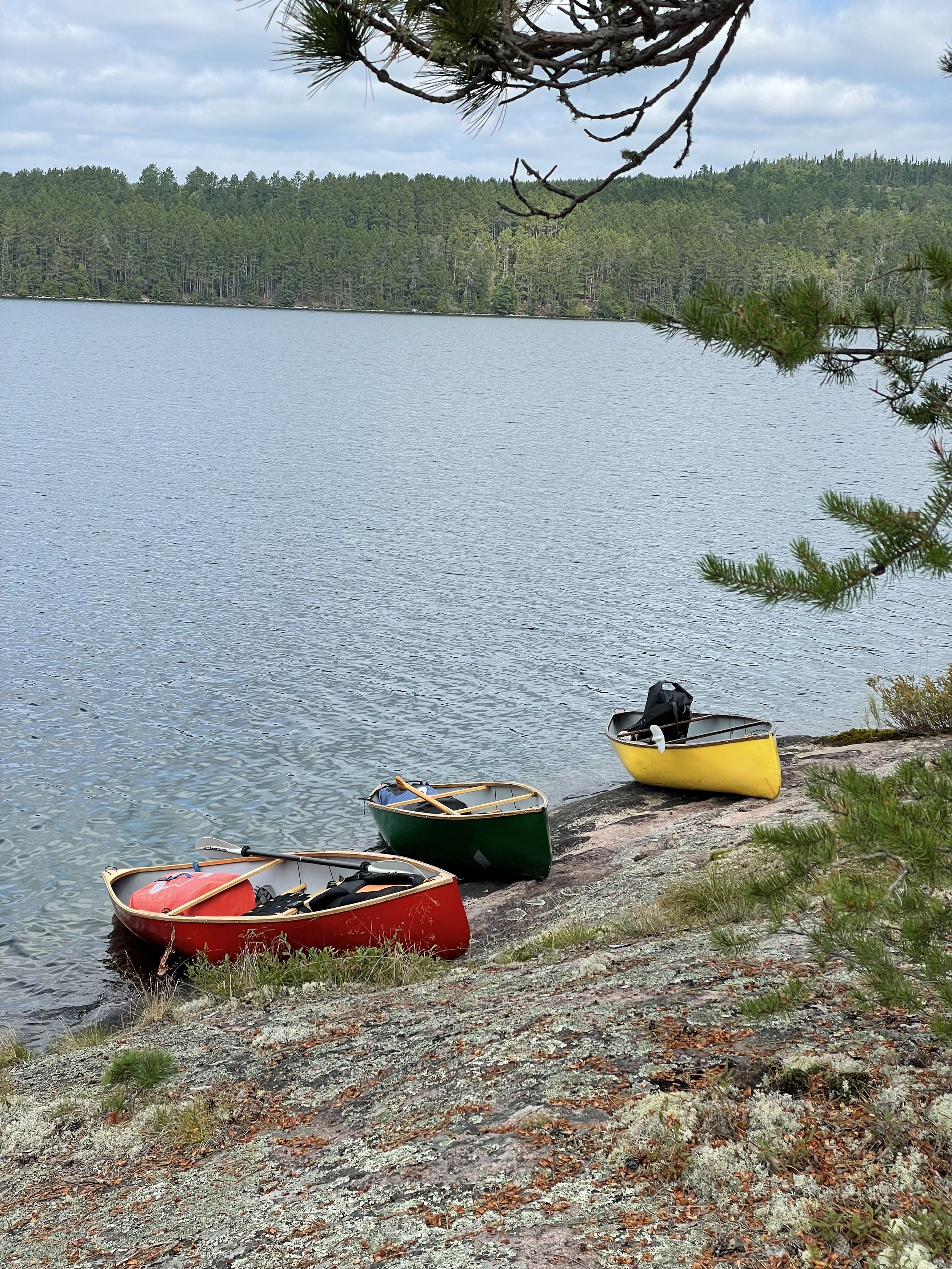 Three colorful canoes, red, green, and yellow, on a rocky lakeshore with a forested background, under a cloudy sky.