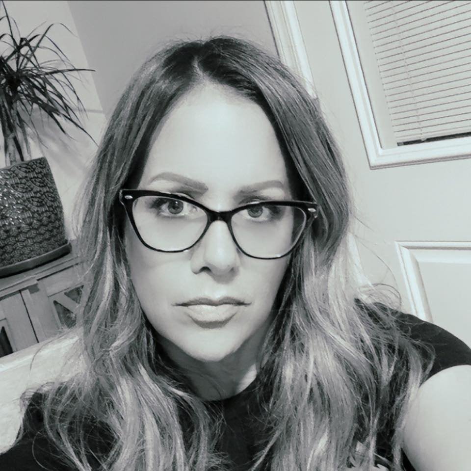 Black and white close-up selfie of a woman with long wavy hair wearing glasses, indoors with a plant and window in the background.