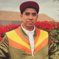 A young man in a graduation cap and gown standing outdoors in front of red flowers and greenery.