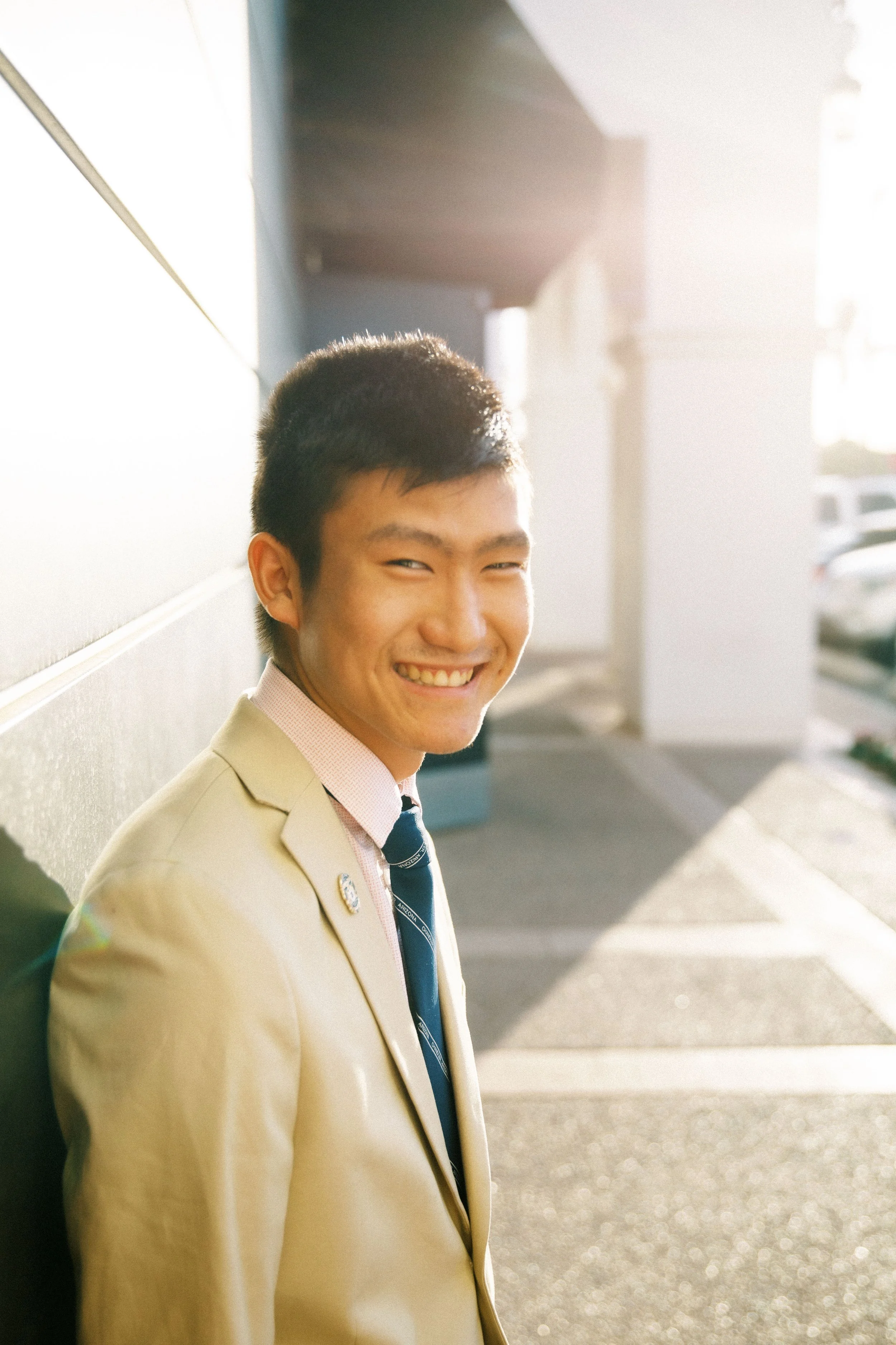 Young man in beige suit smiling outdoors in sunlight near a building.