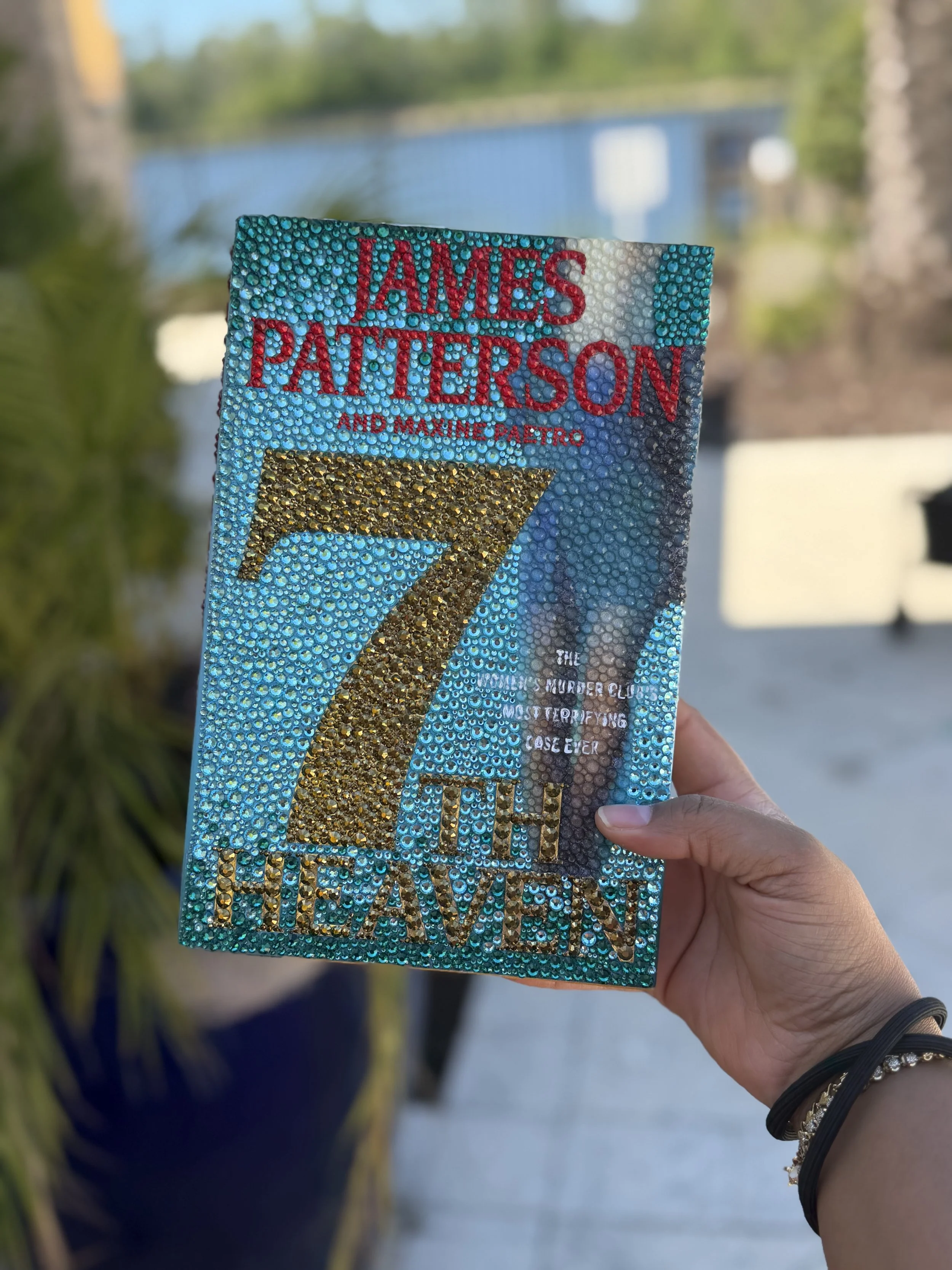 A hand holding a sparkly, decorated campaign sign for James Patterson and Maylene Paletro, with a blue background and gold and red glitter accents, against an outdoor background with trees and a building.