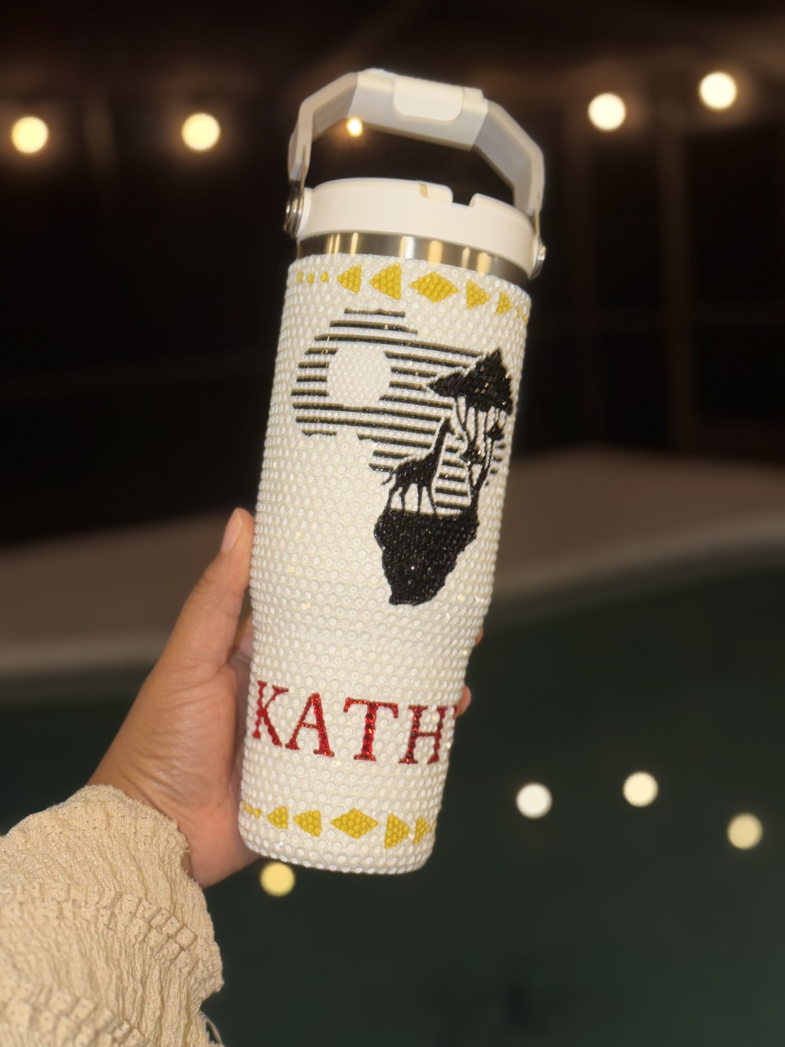 A hand holding a personalized tumbler decorated with rhinestones, featuring the outline of the African continent, a silhouette of a woman, and the name 'KATH' in red letters.
