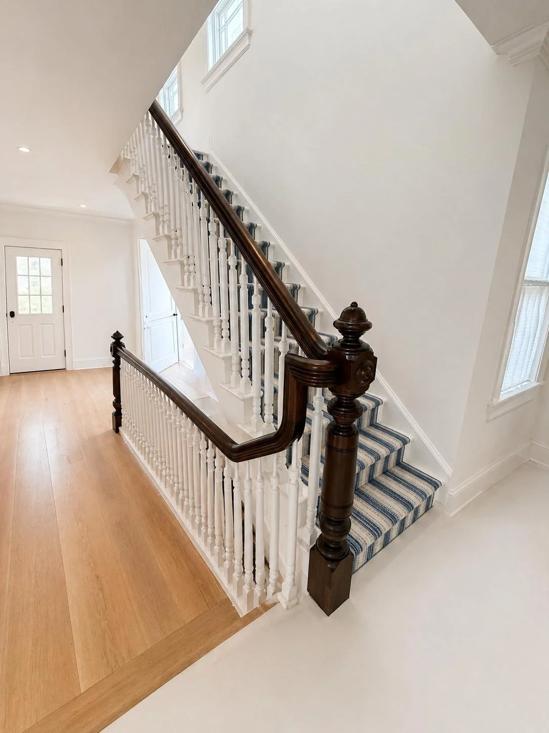 Interior view of a staircase with a dark wooden handrail, white balusters, striped carpet on the stairs, and a hardwood floor at the bottom; white walls with multiple windows, and a door in the background.