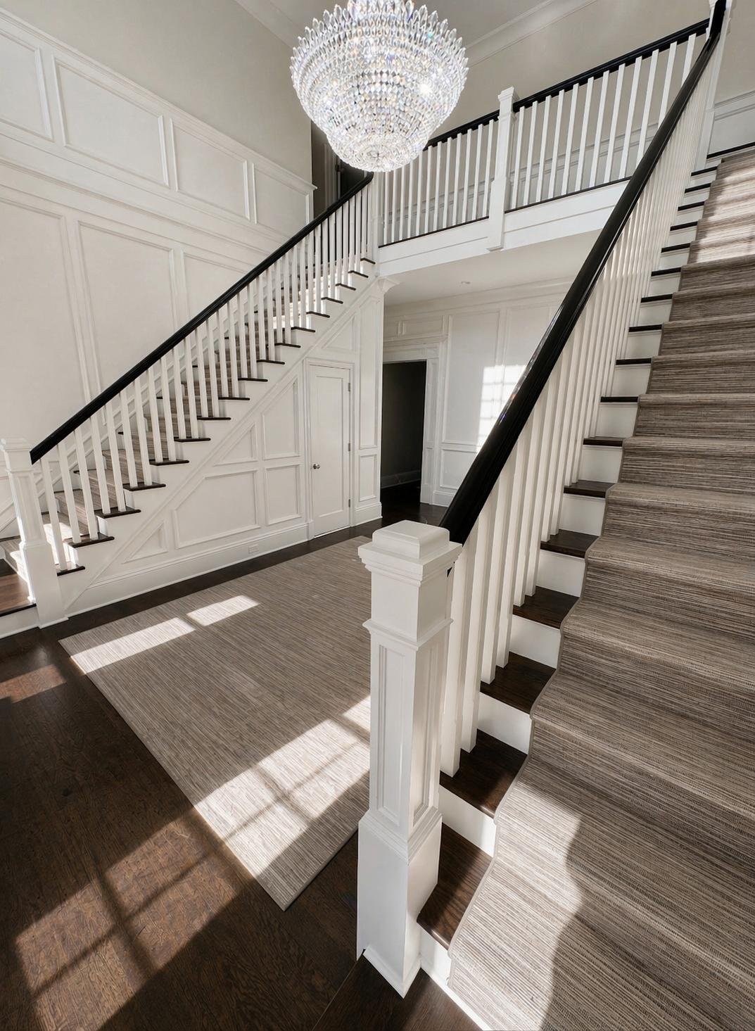 Interior view of a staircase with dark wooden steps, white risers, black handrails, and a beige striped carpet runner. A large, elegant chandelier hangs from the ceiling, and white walls with decorative paneling.