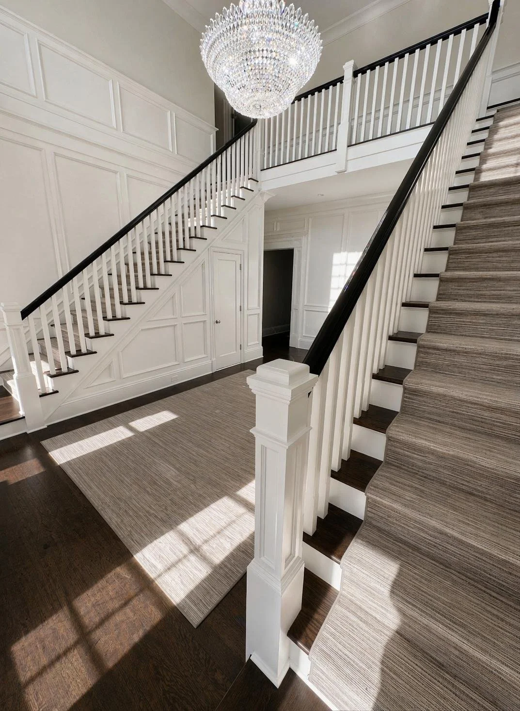 Interior view of a staircase with white balustrades, dark wood handrails, and a beige striped carpet on the stairs, illuminated by sunlight with a large chandelier hanging from the ceiling.